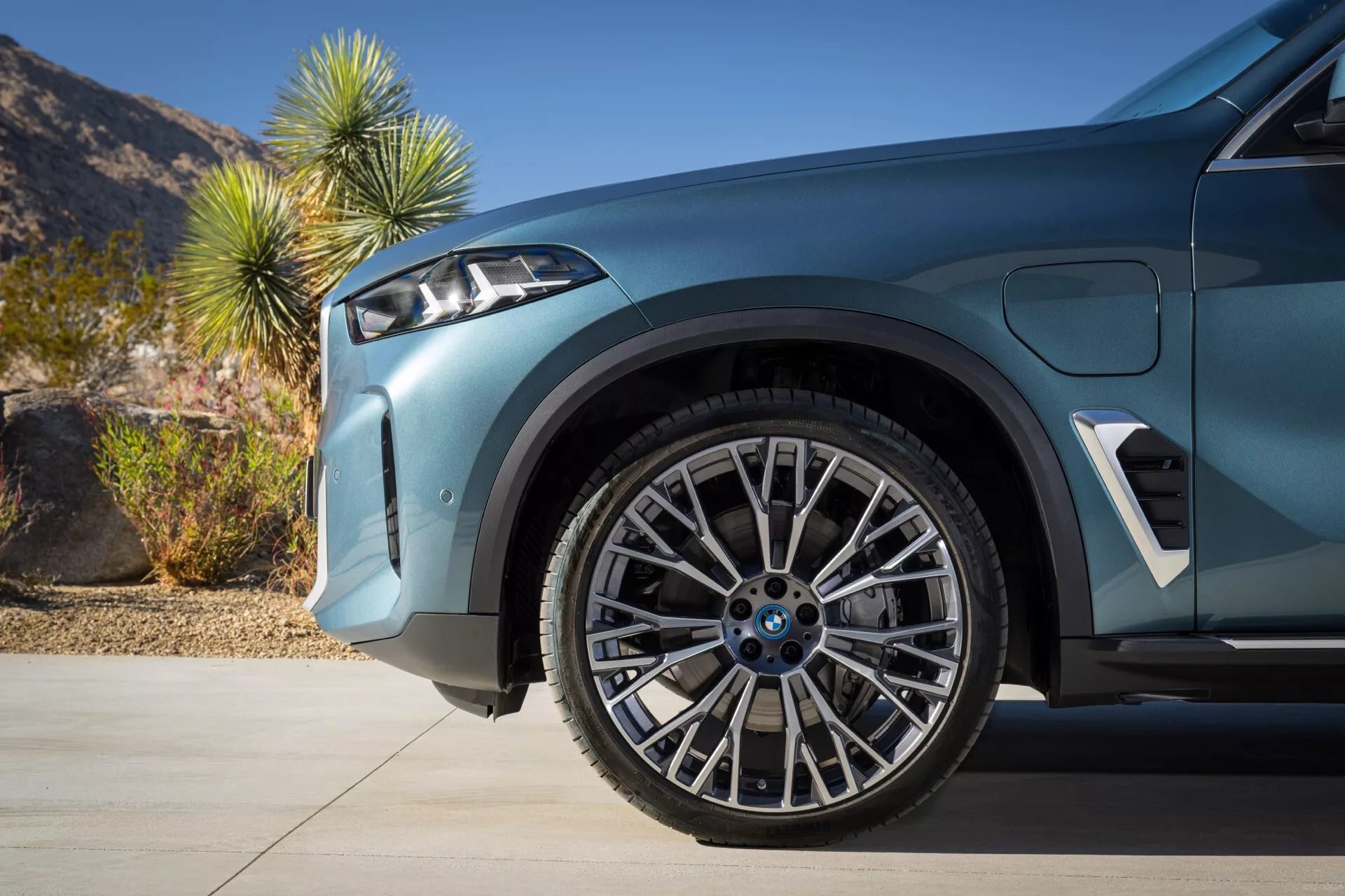 Detajlen posnetek BMW X5 (G05-F95) po osvežitvi v letu 2023, ki poudarja sprednje kolo in blatnik z naprednim vzmetenjem, dvobarvno zvezdasto lito kolo in pnevmatiko z nizkim profilom za zmogljivost, prikazuje športen aerodinamičen dizajn in tehnologijo parkirne asistence ob dnevni puščavski kulisi.