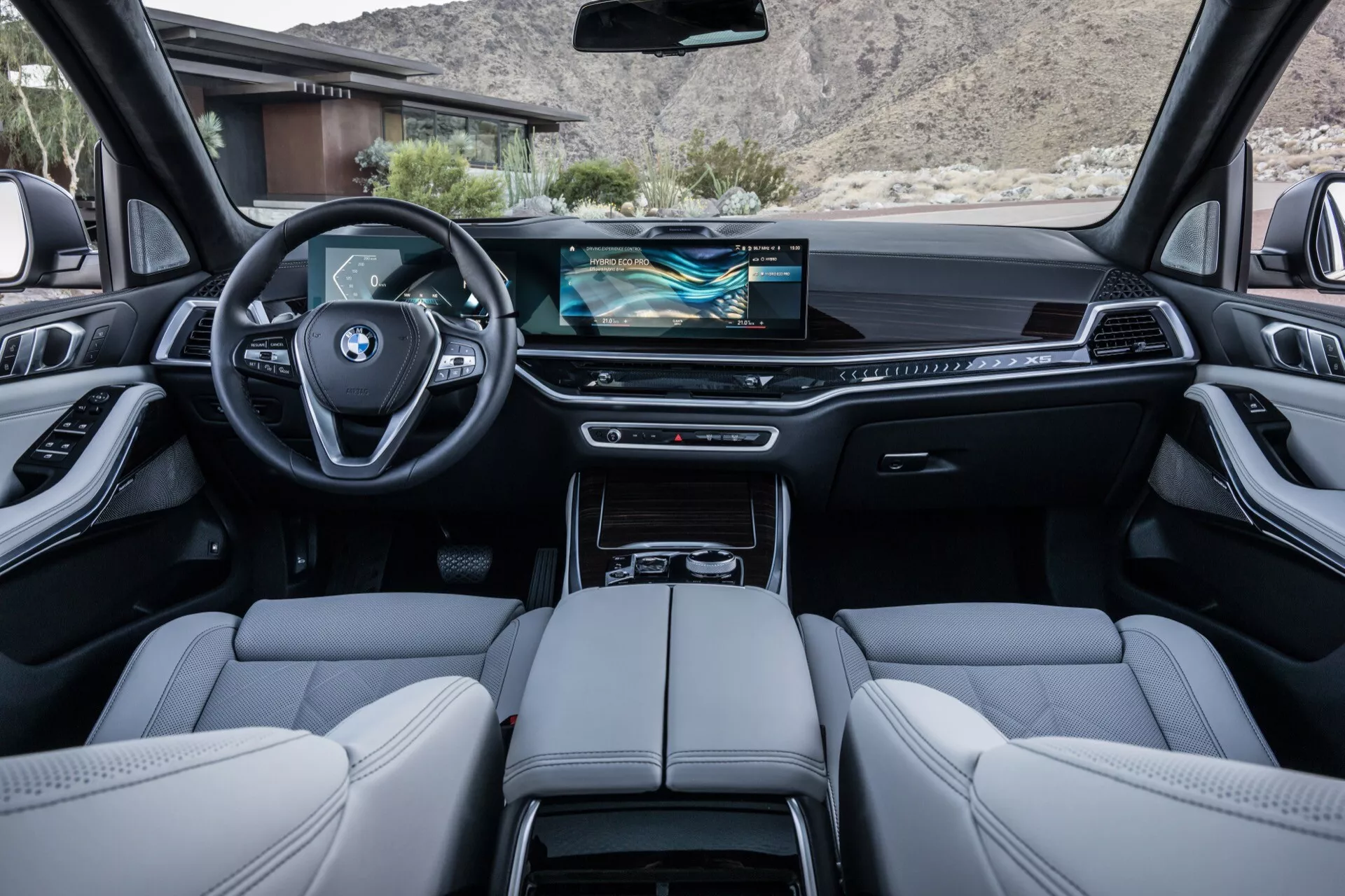 Notranjost BMW X5 2023 (G05 F95 prenova) z zadnjega sedeža s pogledom na armaturno ploščo z zaslonom infozabave širokega zaslona, športnim volanom, oblečenim v usnje, in luksuznimi zaključki, vključno s sedeži iz finega usnja in kovinskimi poudarki