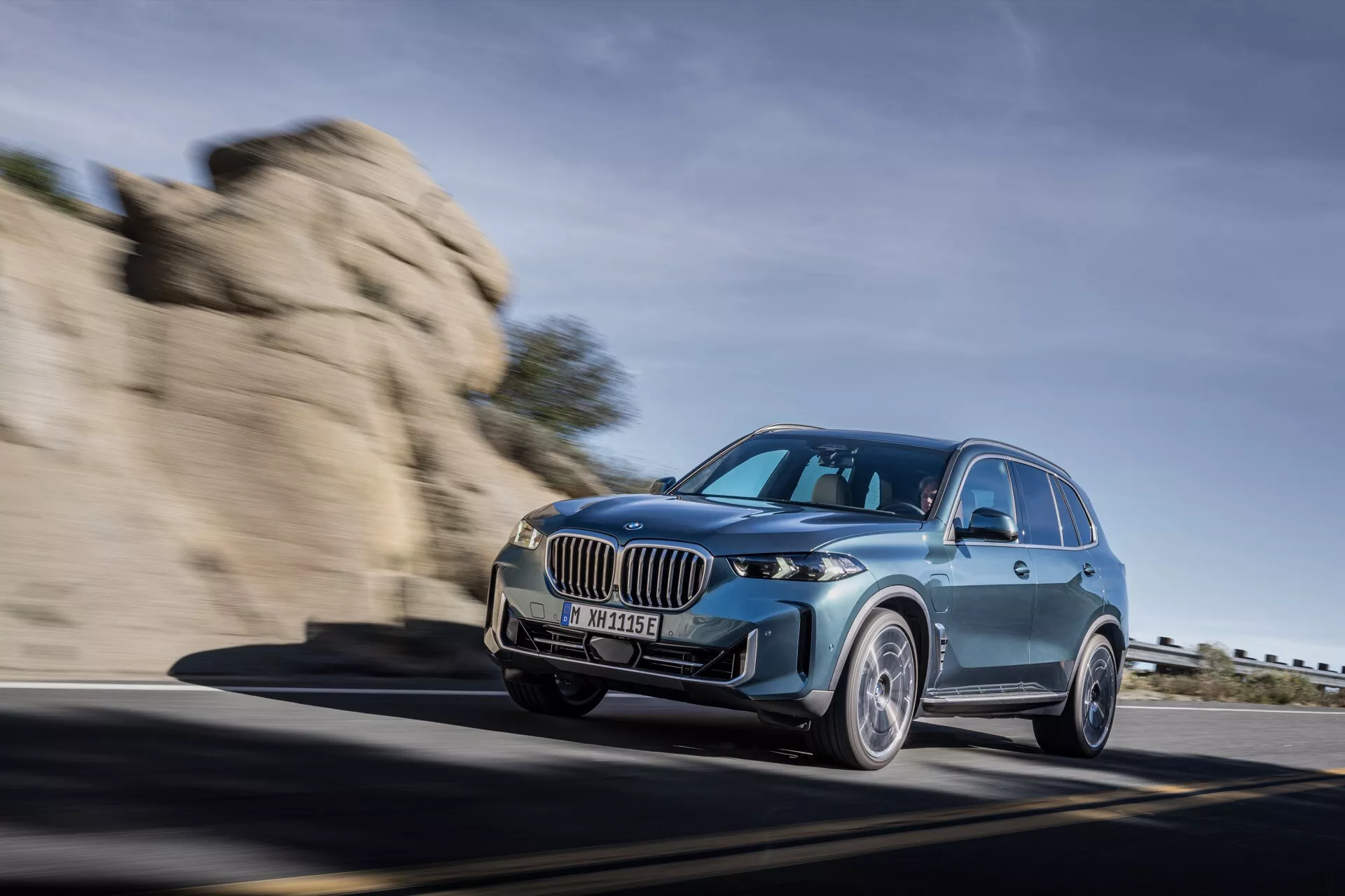 Luksuzni SUV BMW X5 2023 (G05-F95 Facelift) v gibanju, prikazan v dinamični vožnji po gorski cesti z nizkim pogledom spredaj tri četrtine, ki poudarja njegove elegantne LED žaromete, veliko ledvično masko in aerodinamično obliko.