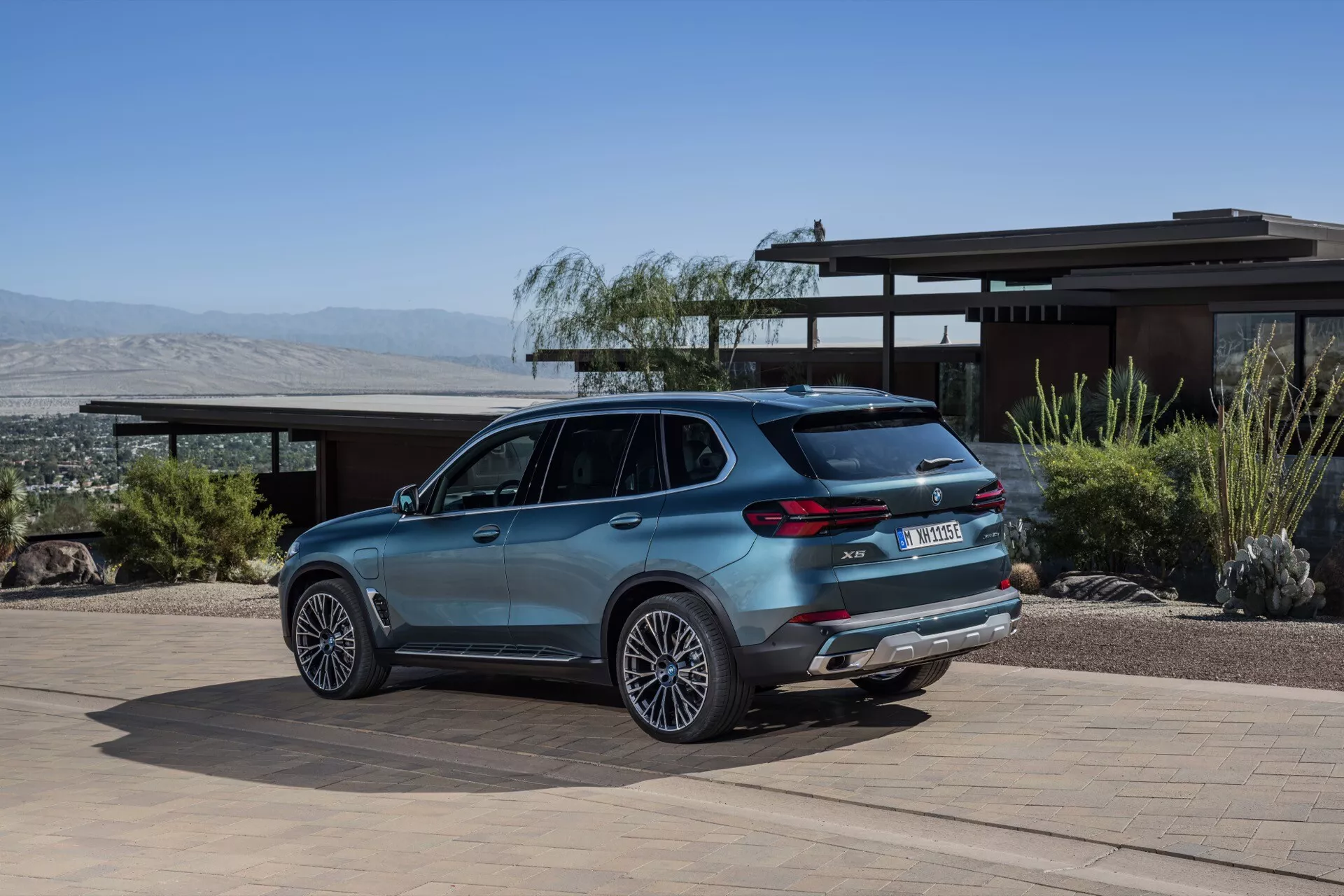 Razkošni SUV BMW X5 2023 (G05-F95 Facelift) parkiran na dovozu stanovanjske hiše z ozadjem puščavske pokrajine in gor, prikazuje lita kolesa, dvojni izpuh, LED zadnje luči, zadnji spojler in strešne nosilce iz kota tri četrtine nazaj.