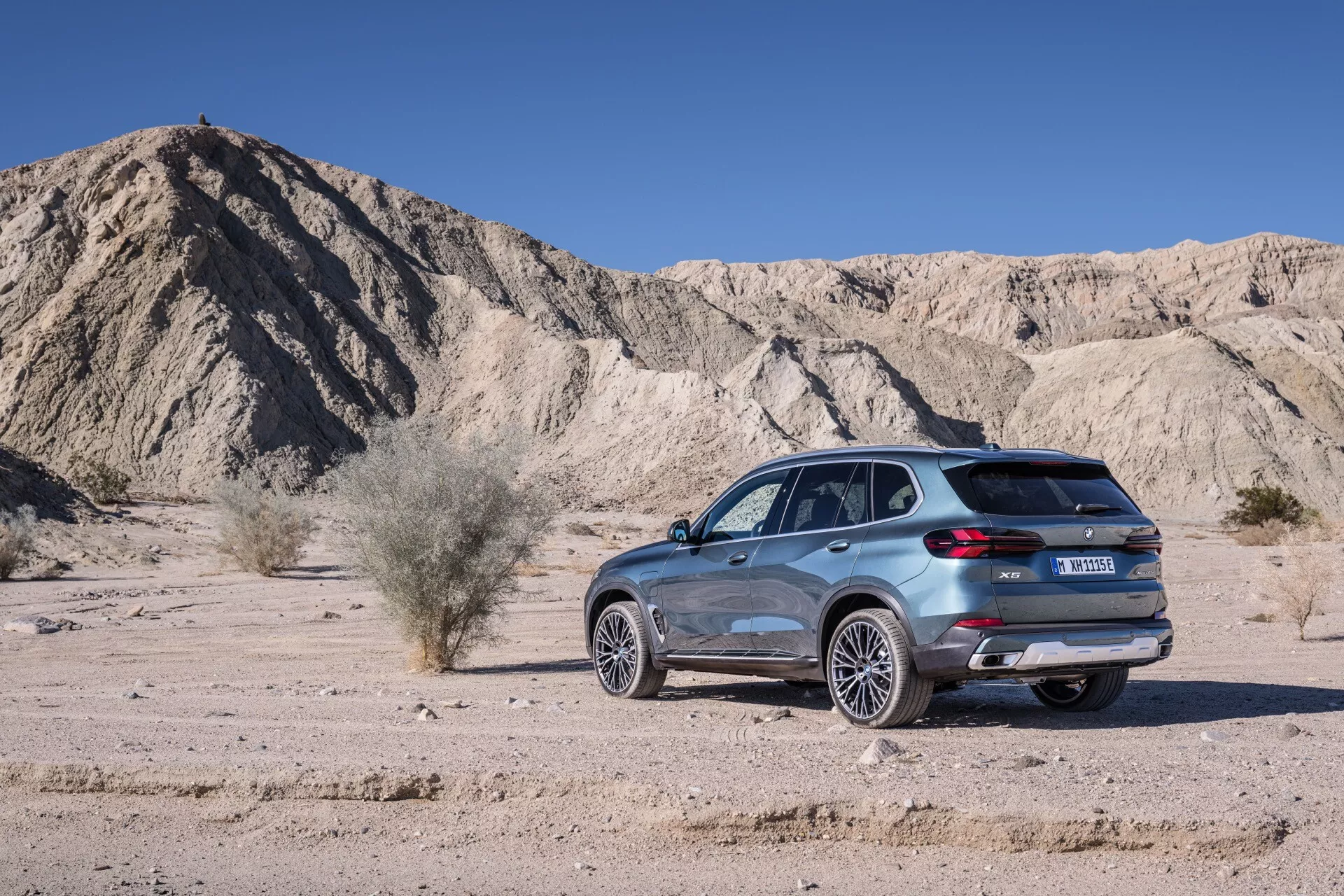 Stranski profil modela BMW X5 (G05-F95) facelift 2023 proti naravnemu puščavskemu pokrajini, z LED razsvetljavo, aerodinamično zasnovo, naprednimi sistemi koles in izpuha ter morebitnimi parkirnimi in vzmetnimi tehnologijami.