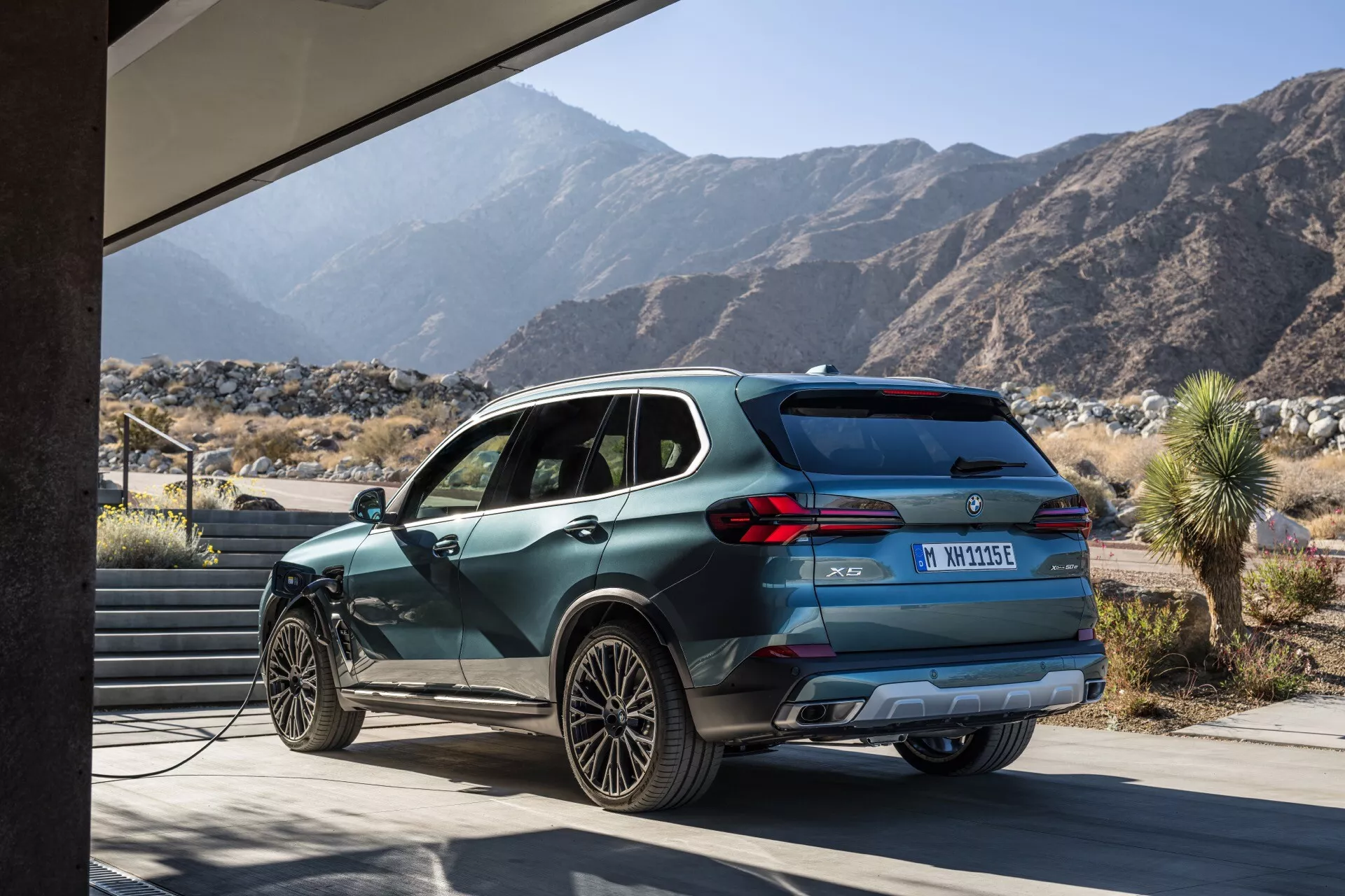 Profilni pogled na BMW X5 2023 (G05-F95-Facelift), ki poudarja njegov aerodinamičen dizajn, sodobno oblikovanje koles in funkcijo polnjenja, proti sodobni strukturi in gorski pokrajini, kar poudarja njegovo ekološko električno ali hibridno naravo in vsestranske zmožnosti v različnih okoljih.