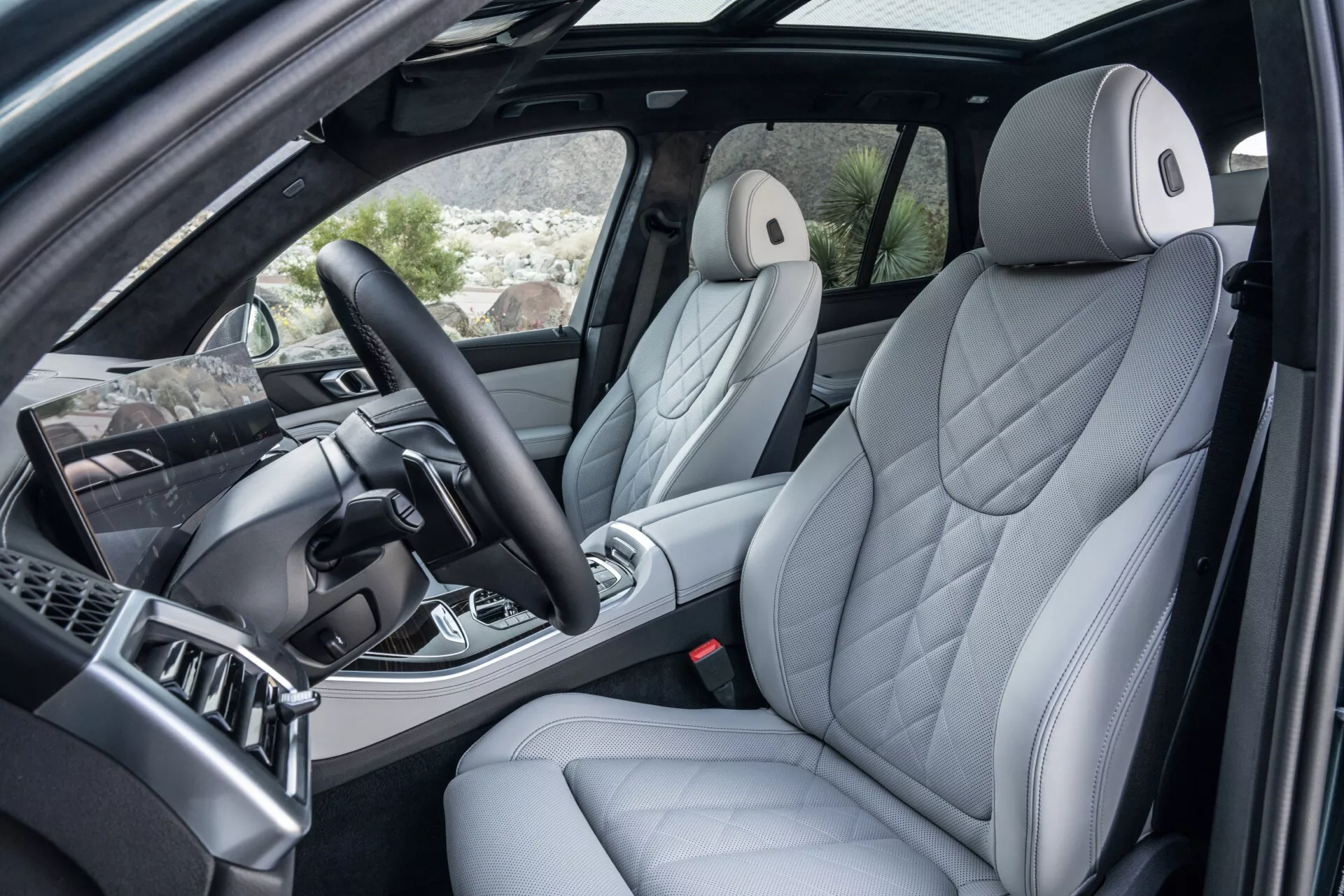 Notranjost BMW X5 2023 (G05 F95 prenova) skozi odprta vrata sovoznika prikazuje razkošen voznikov sedež z ergonomskim prešitim oblazinjenjem, napredna sredinska konzola z ročico prestav in kovinskimi poudarki, večfunkcijski volan in del armaturne plošče z informacijskim sistemom