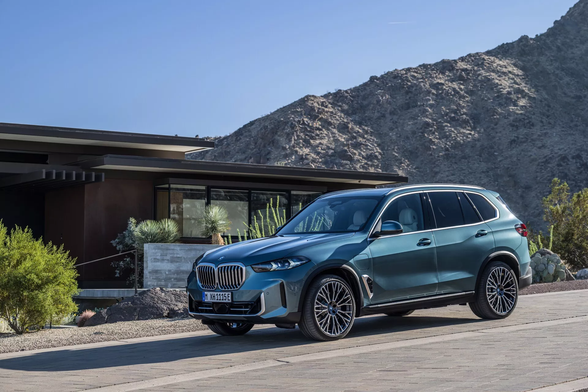 Tričetrtinski sprednji pogled na luksuzni križanec SUV BMW X5 2023 (G05-F95-Facelift) pred prestižno stanovanjsko stavbo z gorami v ozadju, poudarjajoč njegov velik ledvični žar, prilagodljive LED žaromete, večkraka lita platišča, dinamične linije karoserije in napredne značilnosti oblikovanja.