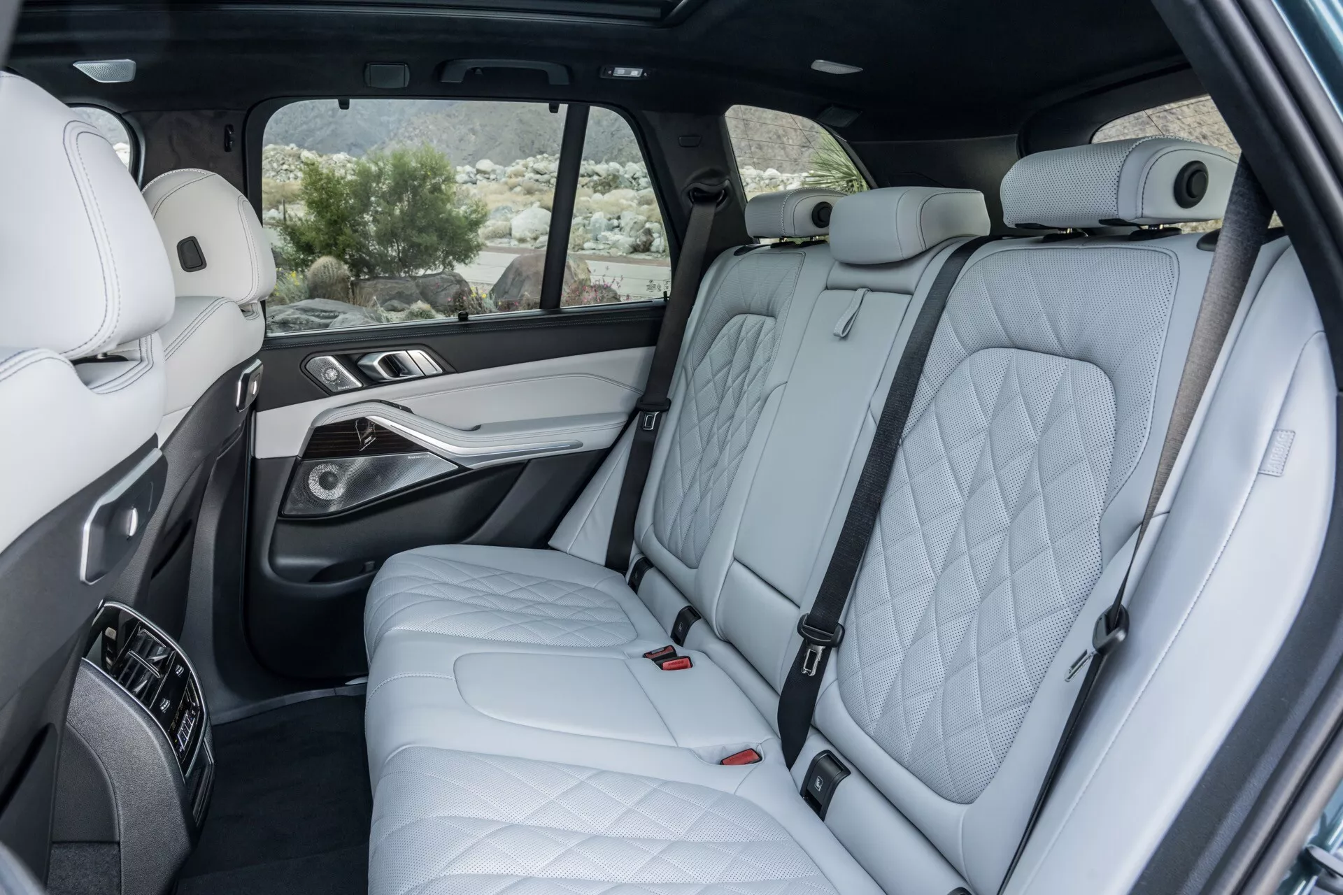 Perspektiva notranjosti BMW X5 2023 (G05-F95-Facelift) z osvetlitvijo razkošnih drugih vrst sedežev s prešito usnje, premijska vrata panel z lesnimi in kovinskimi poudarki, pripomočki sredinske konzole, panoramsko strešno okno, električni kontrolniki zadnjega sedeža in napredne lastnosti vidnosti