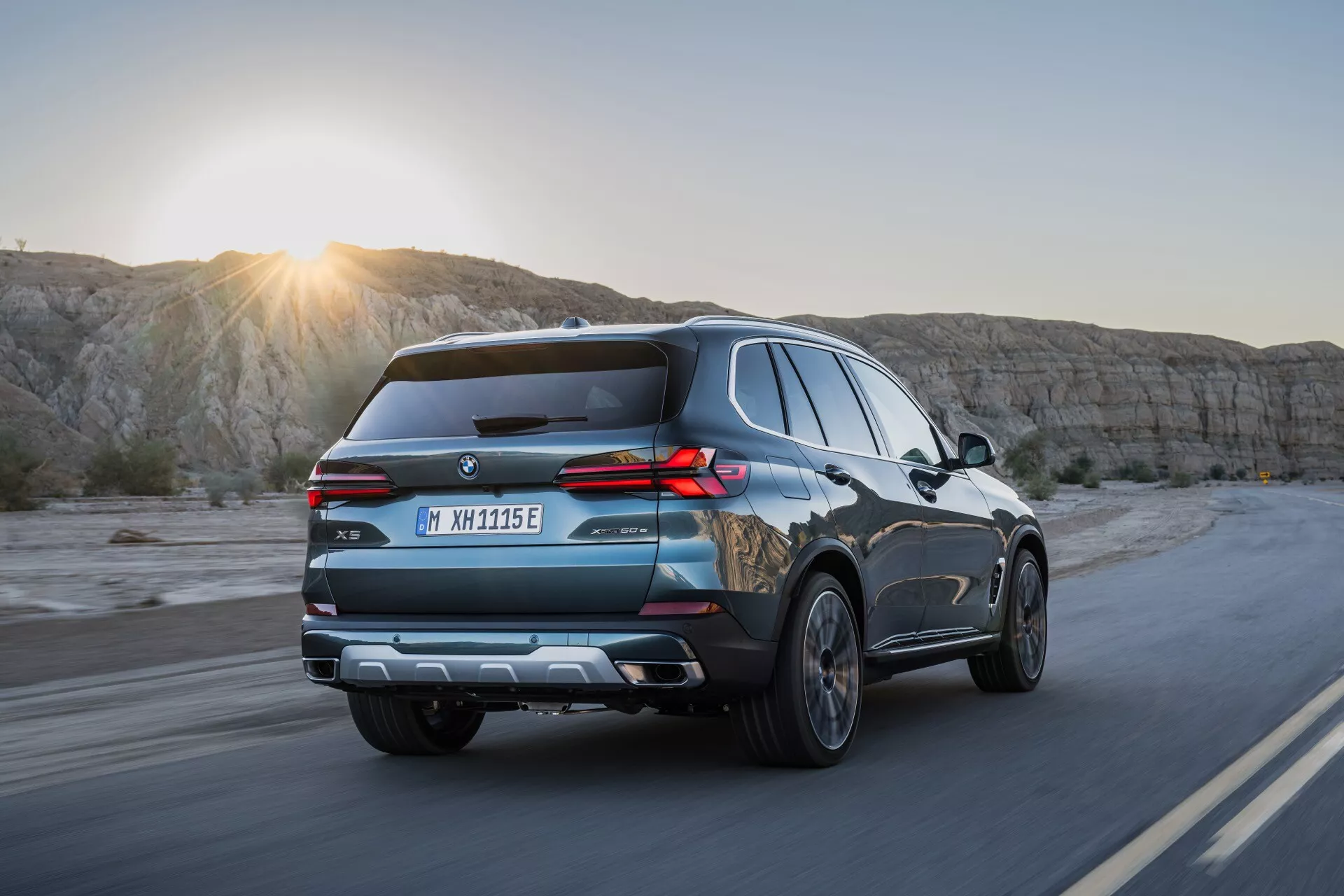 Sodobni SUV BMW X5 2023 (G05-F95-Facelift) v gibanju na slikoviti avtocesti ob sončnem vzhodu ali zahodu, ki zajema dinamični kot zadaj tri četrtine z LED zadnjimi lučmi, strešnimi nosilci in naprednimi tehnološkimi značilnostmi pod nežnim sijajem sonca.