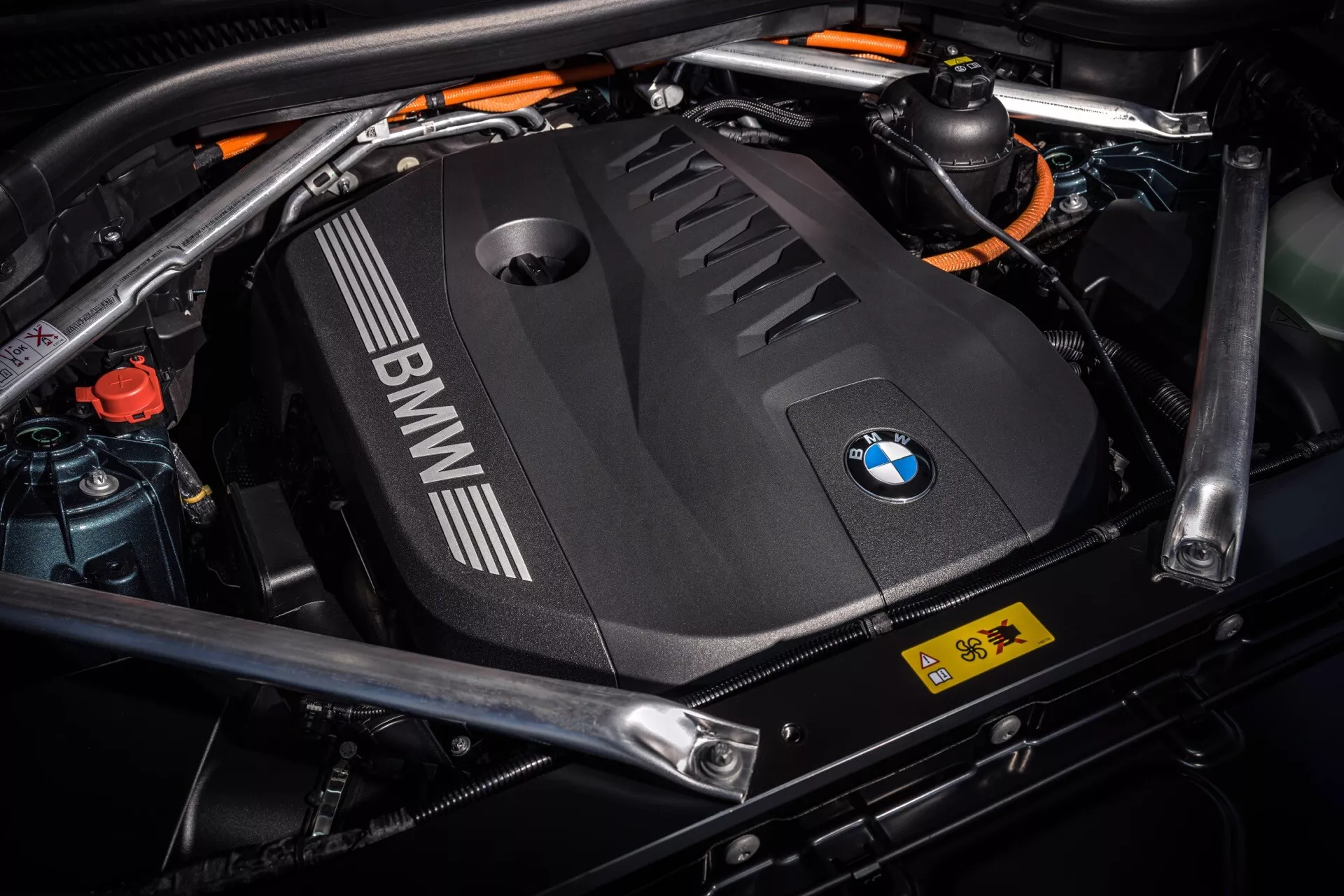 Podroben pogled na motorni prostor BMW X5 2023 (G05-F95-Facelift), ki prikazuje črni pokrov motorja z logotipom BMW, oranžne kable visoke napetosti hibridnega motorja in varnostne oznake, poudarjajoč napredno hibridno tehnologijo in detajle oblikovanja.