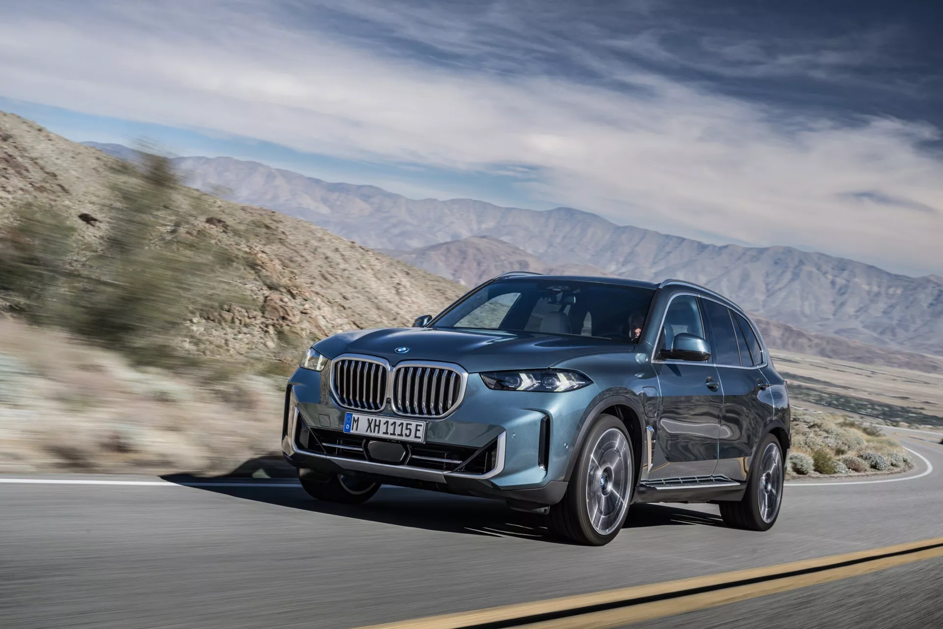 Preoblikovan model BMW X5 2023 (G05-F95) v dinamičnem gibanju, z izstopajočo rešetko, LED žarometi in kovinsko barvo, posnet na sončen dan na gorski cesti