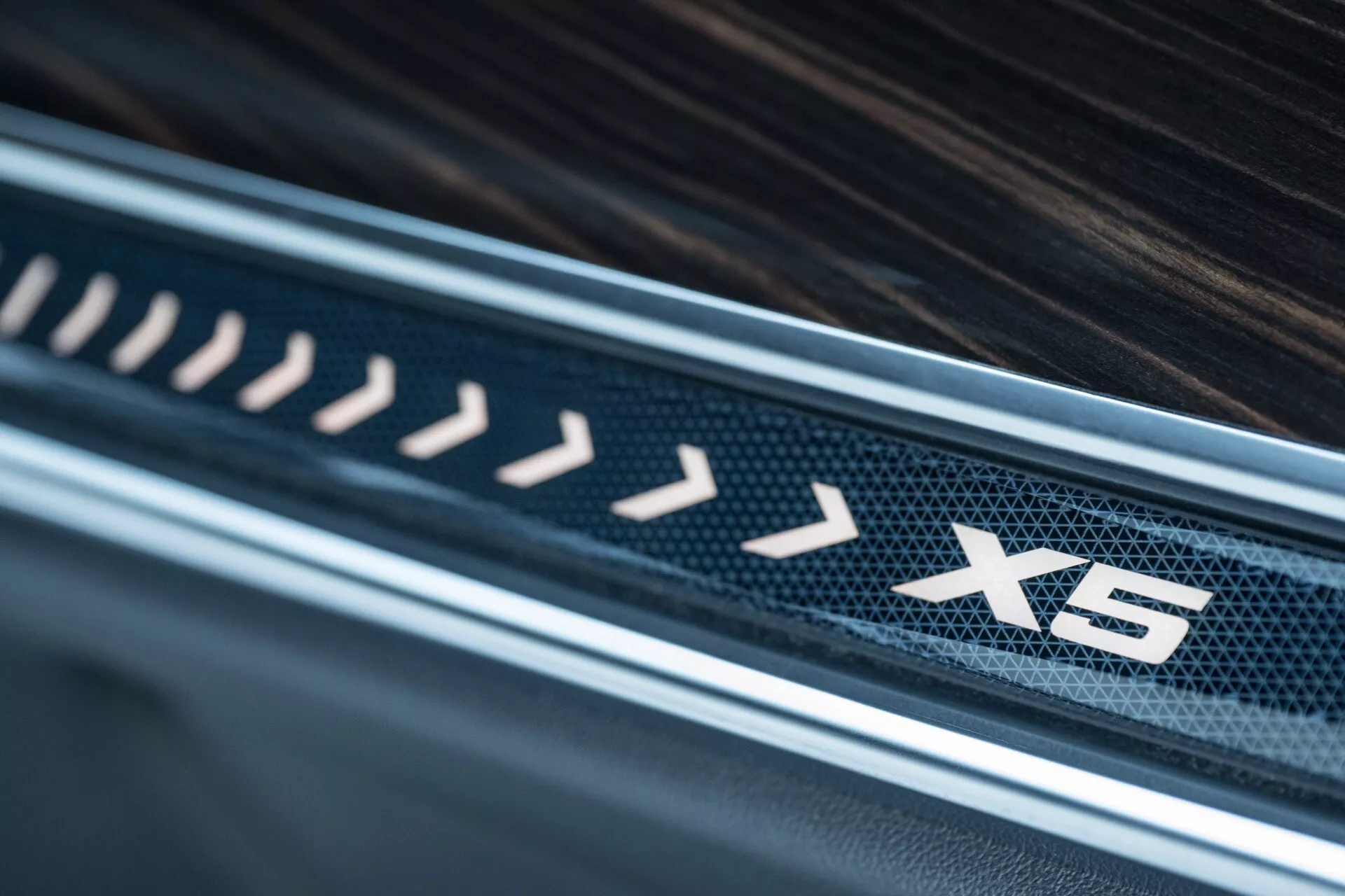 Posnetek notranjosti BMW X5 2023 (G05-F95 facelift) prikazuje podrobnosti luksuznega praga vrat z emblemo 'X5', vzorcem iz ogljikovih vlaken in lesketom, ki poudarja visokokakovostne materiale in oblikovanje
