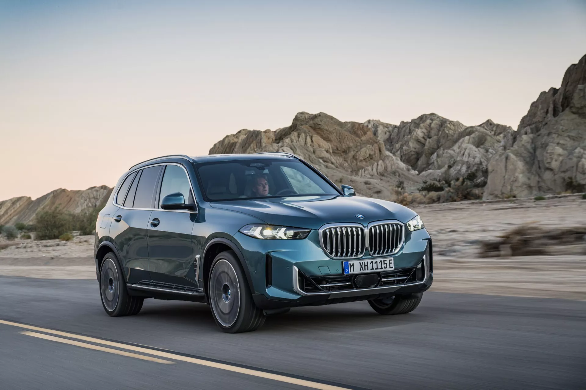 BMW X5 2023 (G05-F95-Facelift) v gibanju na puščavski cesti, prikazuje sprednjo rešetko, LED žaromete, večkraka kolesa in modro-zeleno barvo iz treh četrtin pogleda spredaj v poznem popoldnevu