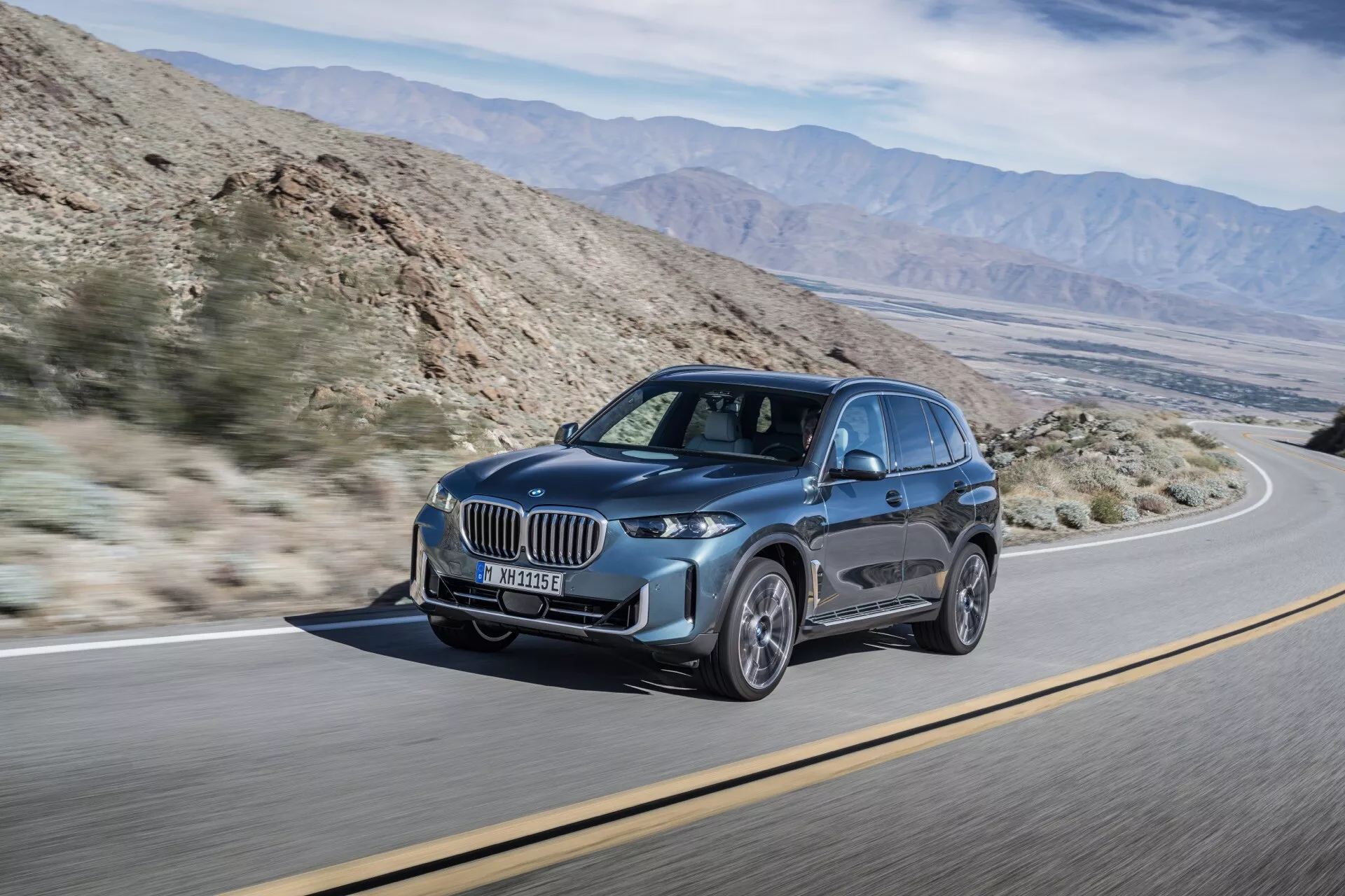 Luksuzni SUV BMW X5 2023 (preoblikovanje G05-F95) v dinamičnem gibanju, prikazuje svoj sodoben dizajn z izstopajočo ledvičasto masko, elegantnimi LED žarometi z dnevnimi lučmi, velikimi liti kolesi z večkrakim designom, in kiparsko aerodinamičnimi linijami karoserije na ozadju, ki spominja na puščavsko pokrajino z gorskimi verigami, poudarja značilnosti kot so napredni sistem razsvetljave in varnostna tehnologija.