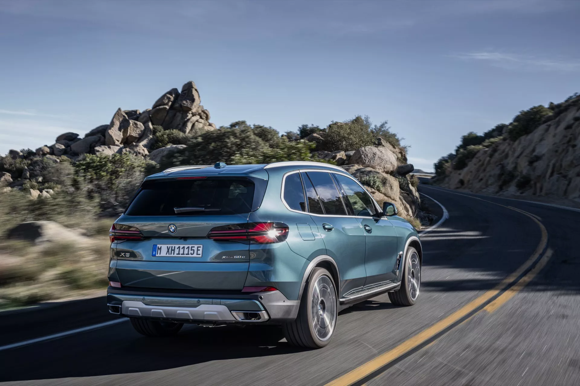 BMW X5 2023 (preoblikovanje G05-F95) v gibanju, posnet z zadnjega tričetrtinskega pogleda, ki poudarja športno zasnovo SUV, sodobni prtljažnik z elegantnimi zadnjimi lučmi, dvobarvna lita kolesa in dokaze o elektrifikaciji na ovinkasti cesti pod jasno dnevno svetlobo