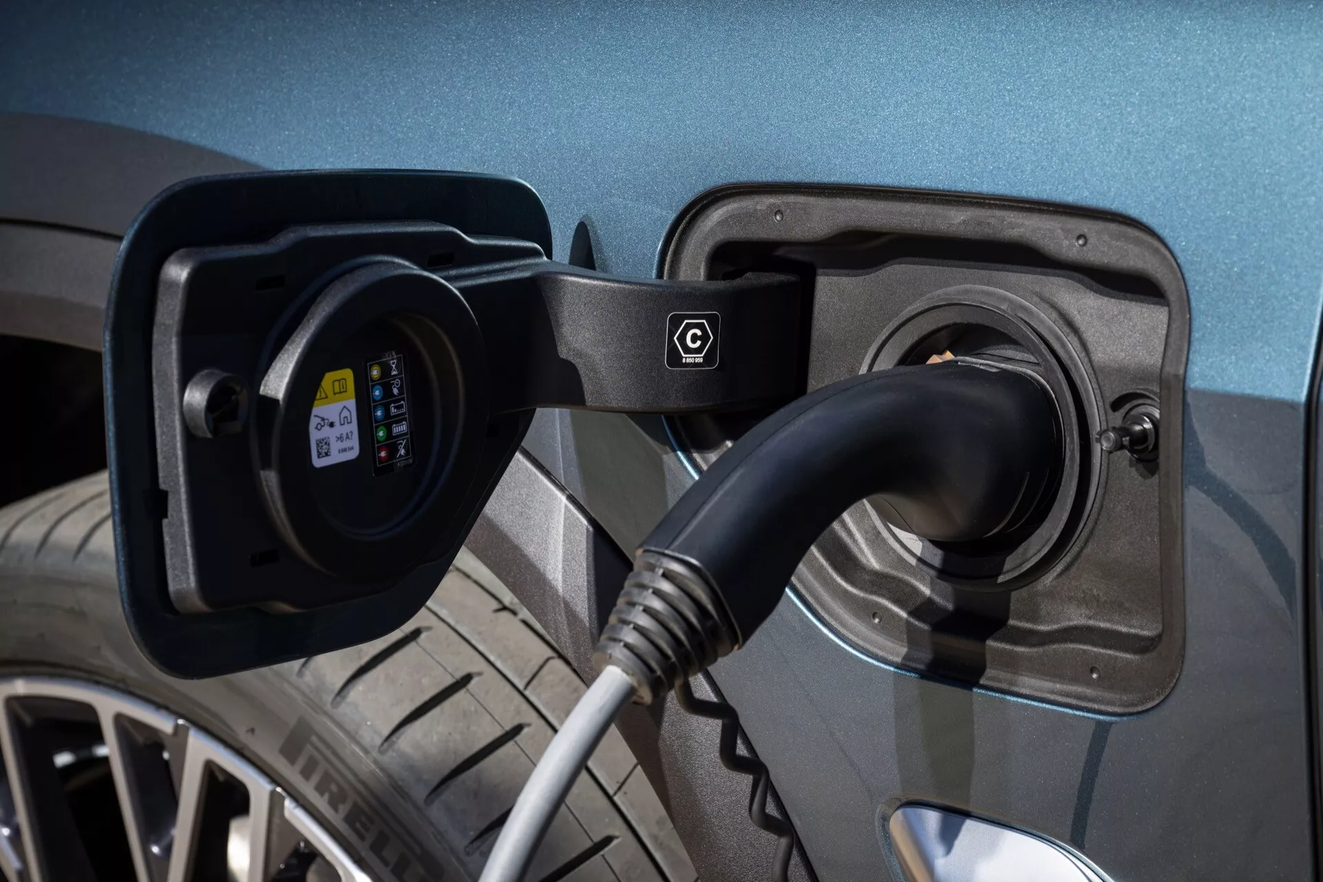 Električno vozilo BMW X5 2023 (G05-F95) z odprtim polnilnim mestom in priključenim polnilnim kablom ter vtičem EV, vidnim indikatorjem statusa polnjenja in podrobnim pogledom na metalno barvo, lito kolo in vzorec tekalne plasti v dobro osvetljenem okolju, ki poudarja moderno tehnologijo trajnostnega prevoza.