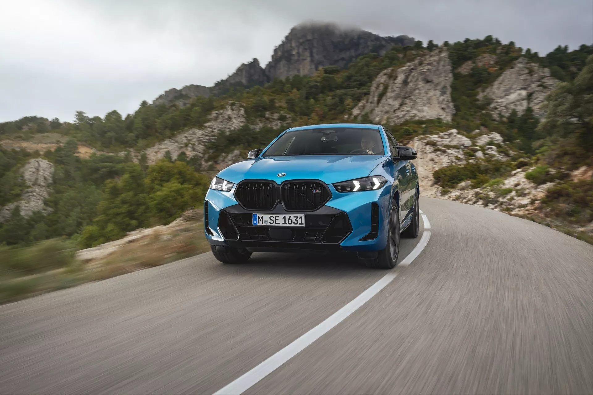 BMW X6 2023 (G06-F96-facelift) v živahni modri barvi, ki predstavlja moderno luksuzno zmogljivost na vijugasti gorski cesti z dinamičnim zamegljevanjem gibanja, naprednimi LED žarometi, elegantnimi oblikovalskimi elementi in športnimi kolesi, na ozadju skalnatega gorskega terena.