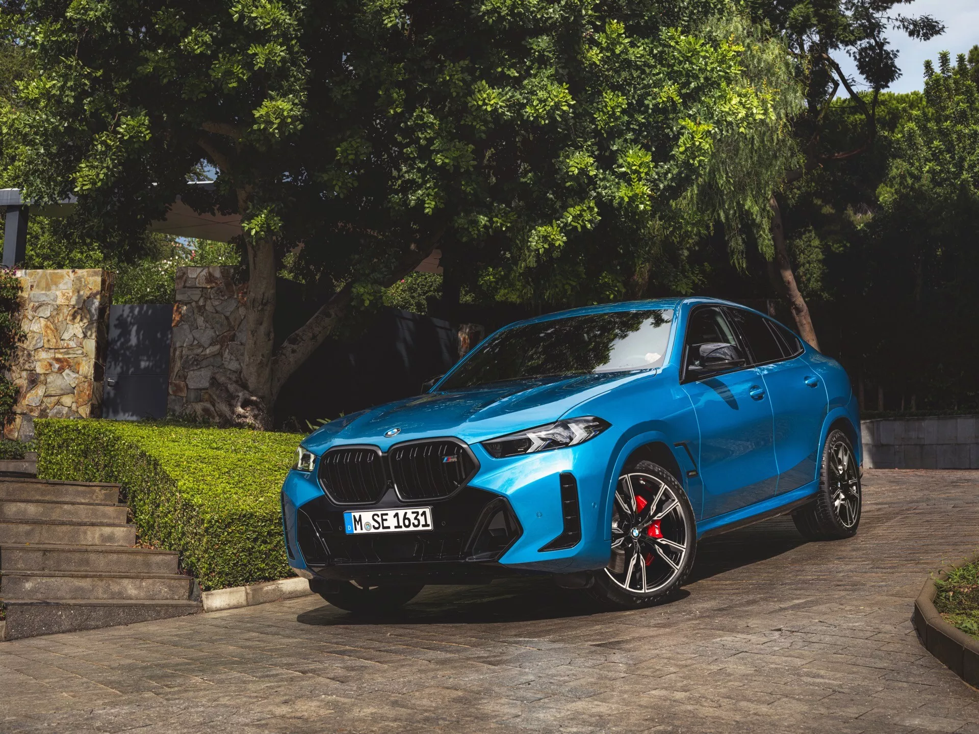 BMW X6 2023 (G06-F96-Facelift) v modri barvi, parkiran pod kotom, prikazuje svojo sprednjo fasado, ledvičasto obliko rešetke, LED ali laserska prednja luči, športna lita kolesa z rdečimi zavornimi čeljusti, konturirane linije pokrova in aerodinamična stranska ogledala z integriranimi smerniki, posneto v naravni dnevni svetlobi z zelenjem in arhitekturnimi stopnicami v ozadju