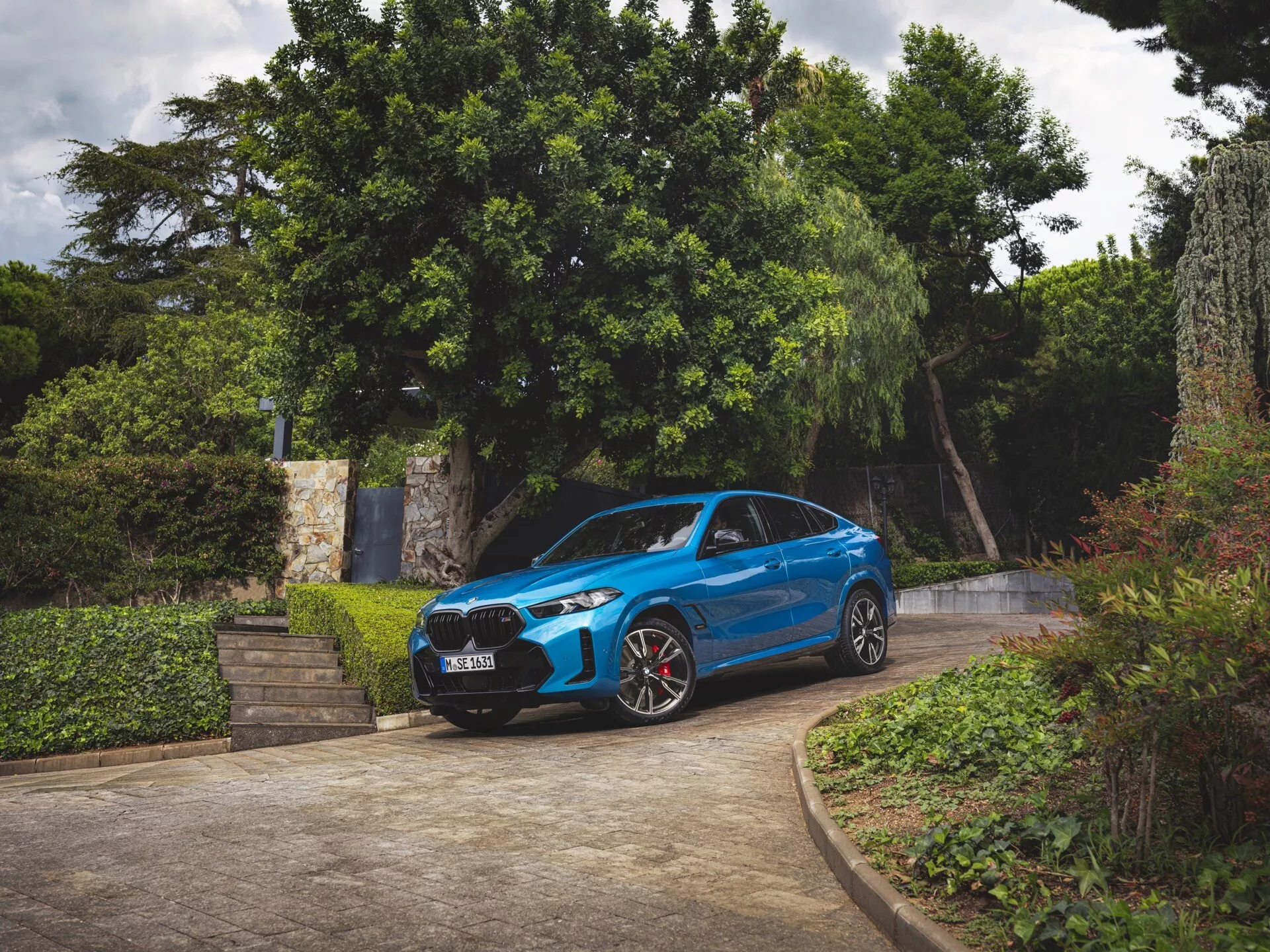 BMW X6 2023 (G06-F96-Preoblikovanje) v modri barvi, pogled spredaj treh četrtin, parkiran na dovozu obdanem z zelenjem, prikazuje sprednji odbijač, rešetko, pokrov motorja, vetrobransko steklo, profil voznikove strani, LED žaromete, športna kolesa z rdečimi zavornimi čeljustmi in aerodinamično obliko