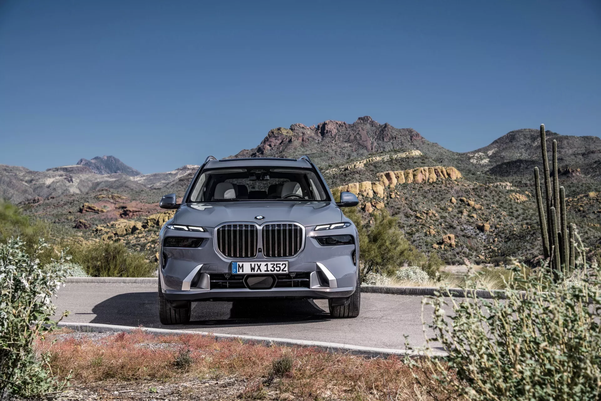 Luksuzni SUV BMW X7 2022 (G07-Facelift), ki prikazuje sodobno oblikovanje z veliko ledvično rešetko, ozkimi LED žarometi, naprednim sprednjim odbijačem z funkcijami ADAS, kipasto oblikovanim pokrovom motorja in litoželeznimi kolesi, vpadljivo parkirano na cesti ob ozadju puščavske pokrajine in gora pod jasno modrim nebom
