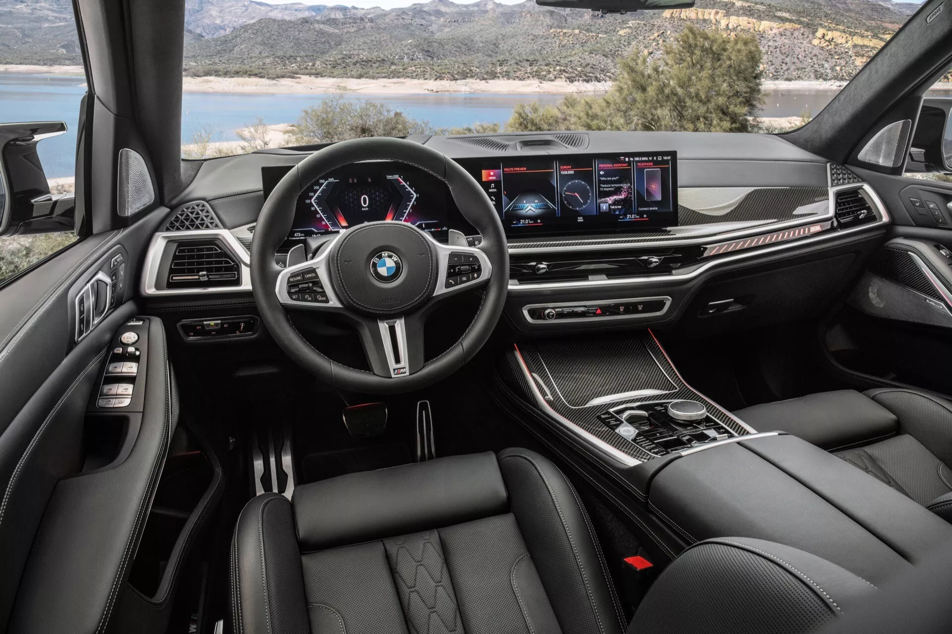 Pogled na notranjost BMW X7 2022 (G07 Facelift) iz perspektive voznika, ki prikazuje multifunkcijski volan z logotipom BMW, digitalni merilnik hitrosti, širokozaslonski zaslon za informacijsko-zabavne vsebine, gumbe za nadzor klime, izbirnik za avtomatski menjalnik, luksuzne materiale, športne in udobne sedeže ter ambientalno razsvetljavo