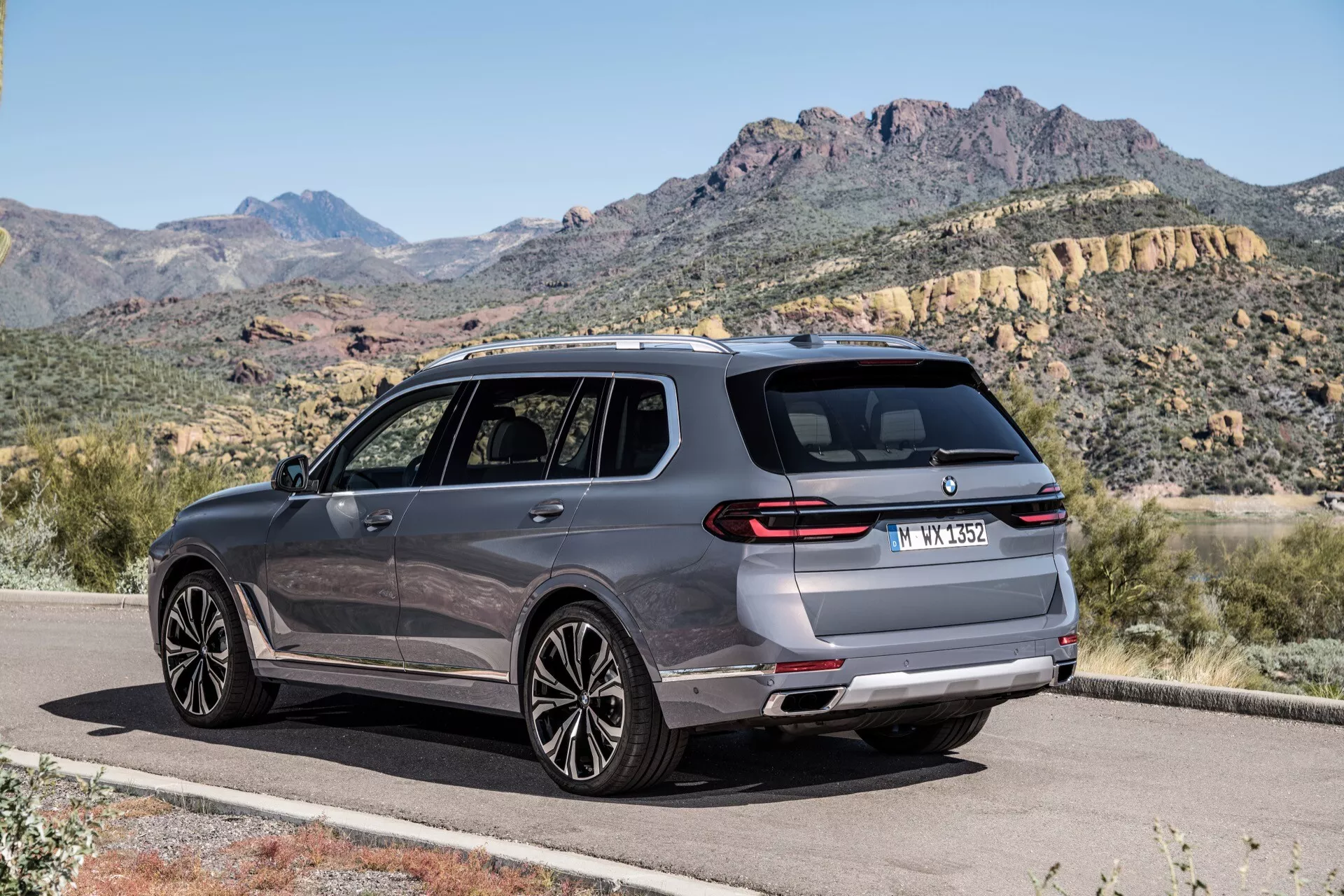 Moderen SUV BMW X7 2022 (G07 Facelift) v metalni barvi na tlakovani cesti proti gorski pokrajini, prikazuje profil zadnjega dela in strani iz treh četrtin, z vidnimi zadnjimi lučmi, zadnjim odbijačem, parkirnimi senzorji, dvojnimi izpušnimi cevmi, platišči iz zlitine z večkrakim dizajnom, okni obrobljenimi s kromom, stranskimi ogledali z integriranimi smerniki in strešnimi nosilci