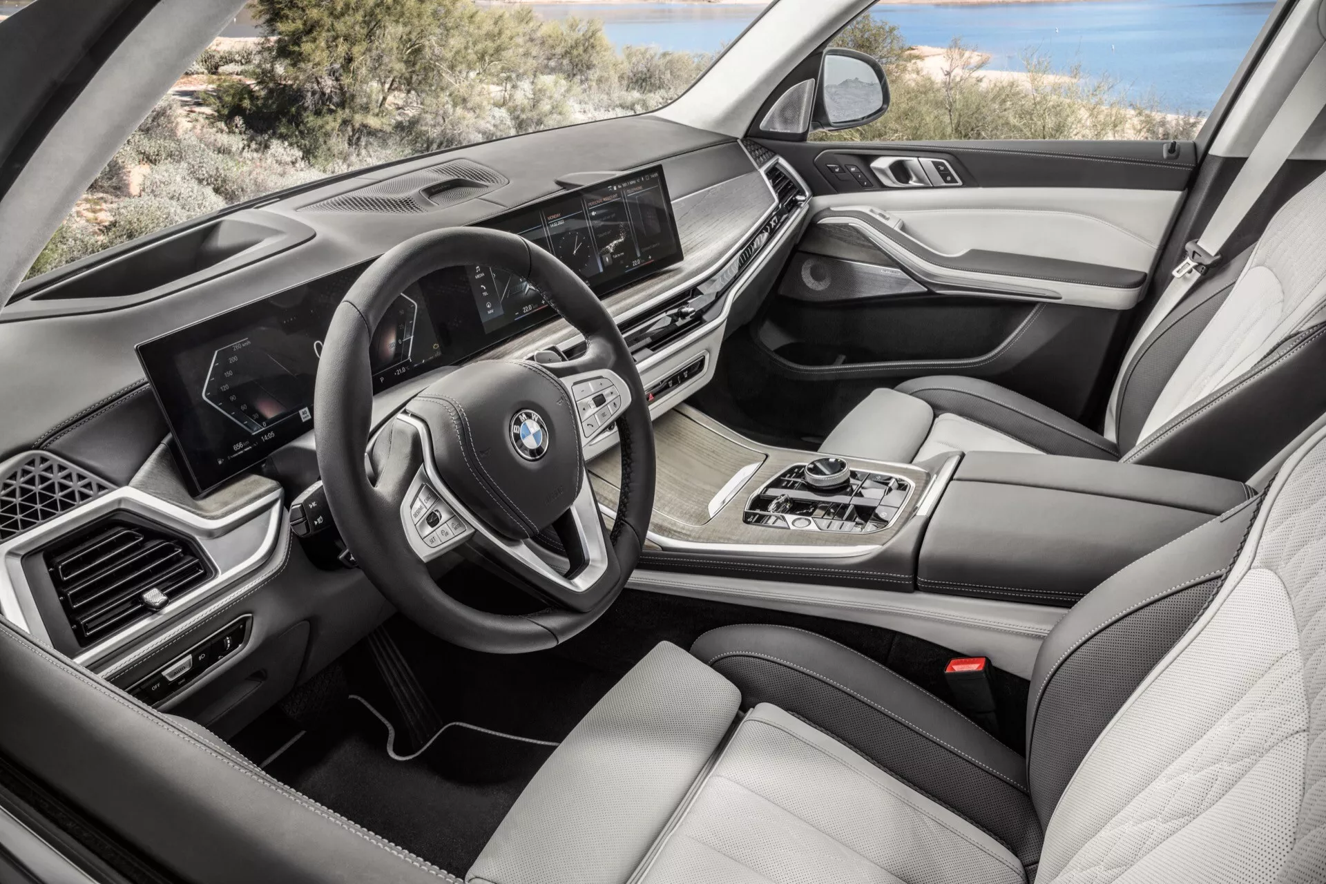 Pogled na notranjost BMW X7 2022 (G07-Facelift) s strani potnika, ki poudarja armaturno ploščo z zaslonom za informacije in zabavo, multifunkcijski volan, avtomatski menjalnik, nadzor multimedijskih naprav iDrive, dvoconsko klimatsko napravo, luksuzne usnjene sedeže, lesene in kovinske poudarke ter digitalni merilnik.