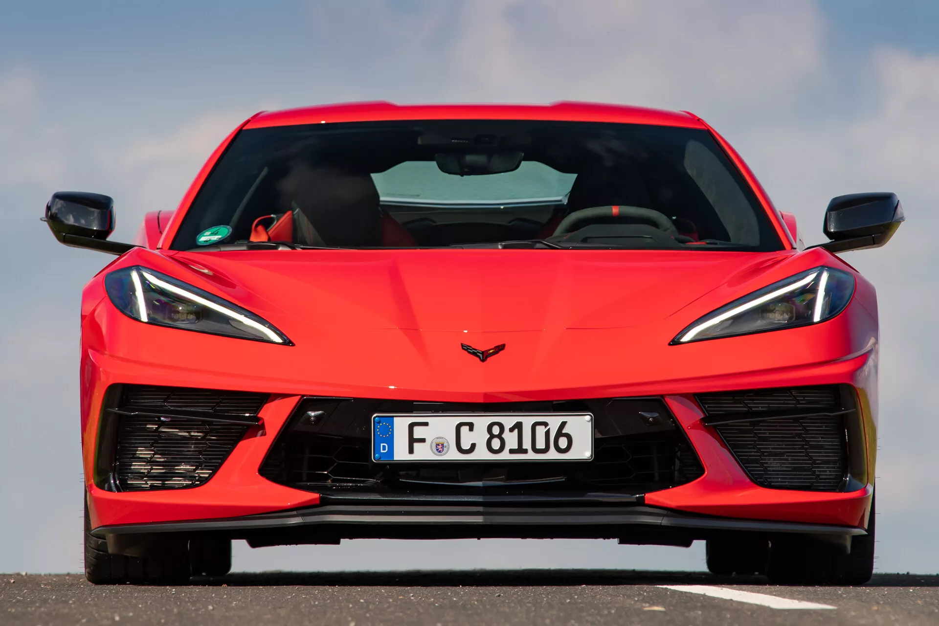 Frontalni pogled na rdeč športni avtomobil Chevrolet Corvette 2021 (C8), posnet pod kotom iz višine oči, ki prikazuje simetrični dizajn avtomobila. Slika poudarja podrobnosti sprednjega dela vozila, vključno z ostrimi, kotnimi LED žarometi, edinstveno mrežico z majhnimi potrebami po hlajenju in velikimi zračniki na spodnjem odbijaču. Emblem Corvette je jasno viden na pokrovu motorja. Ta bližnji posnetek zajame sodoben dizajn in napredne tehnološke elemente avtomobila.