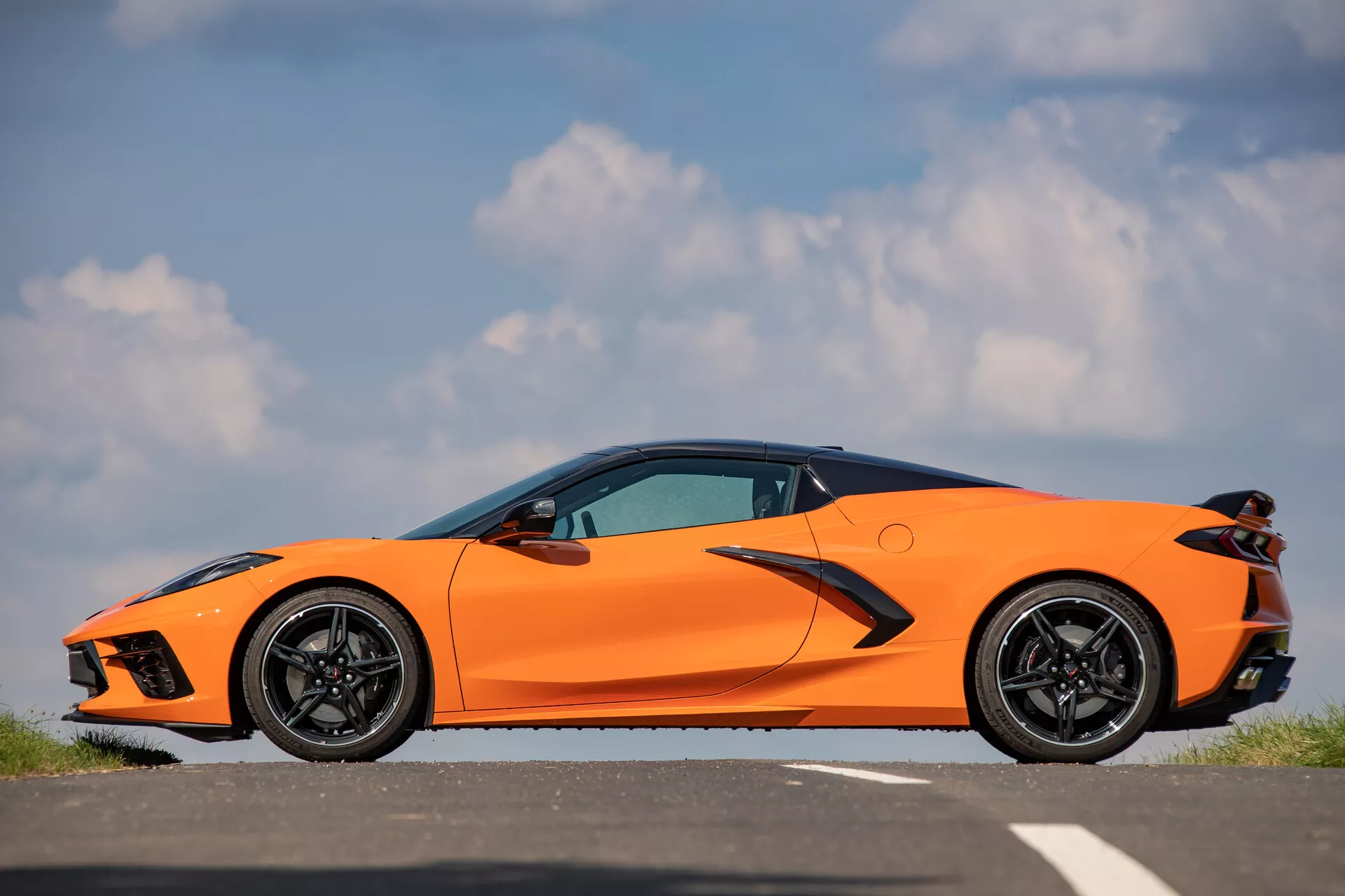 Stranski profil Chevrolet Corvette (C8) 2021 v svetlo oranžni barvi na asfaltni površini, prikazujejo aerodinamično oblikovano sprednjo stran z ostrimi LED žarometi, velikimi zračnimi vstopniki, elegantno silhueto z izrazito značilno linijo, poravnane kljuke vrat, namensko oblikovane stranske krilce, velika črna športna platišča z nizkoprofilnimi zmogljivimi pnevmatikami, izjemno zmogljive zavorne čeljusti in delno vidne kotne zadnje luči proti jasnemu nebu.