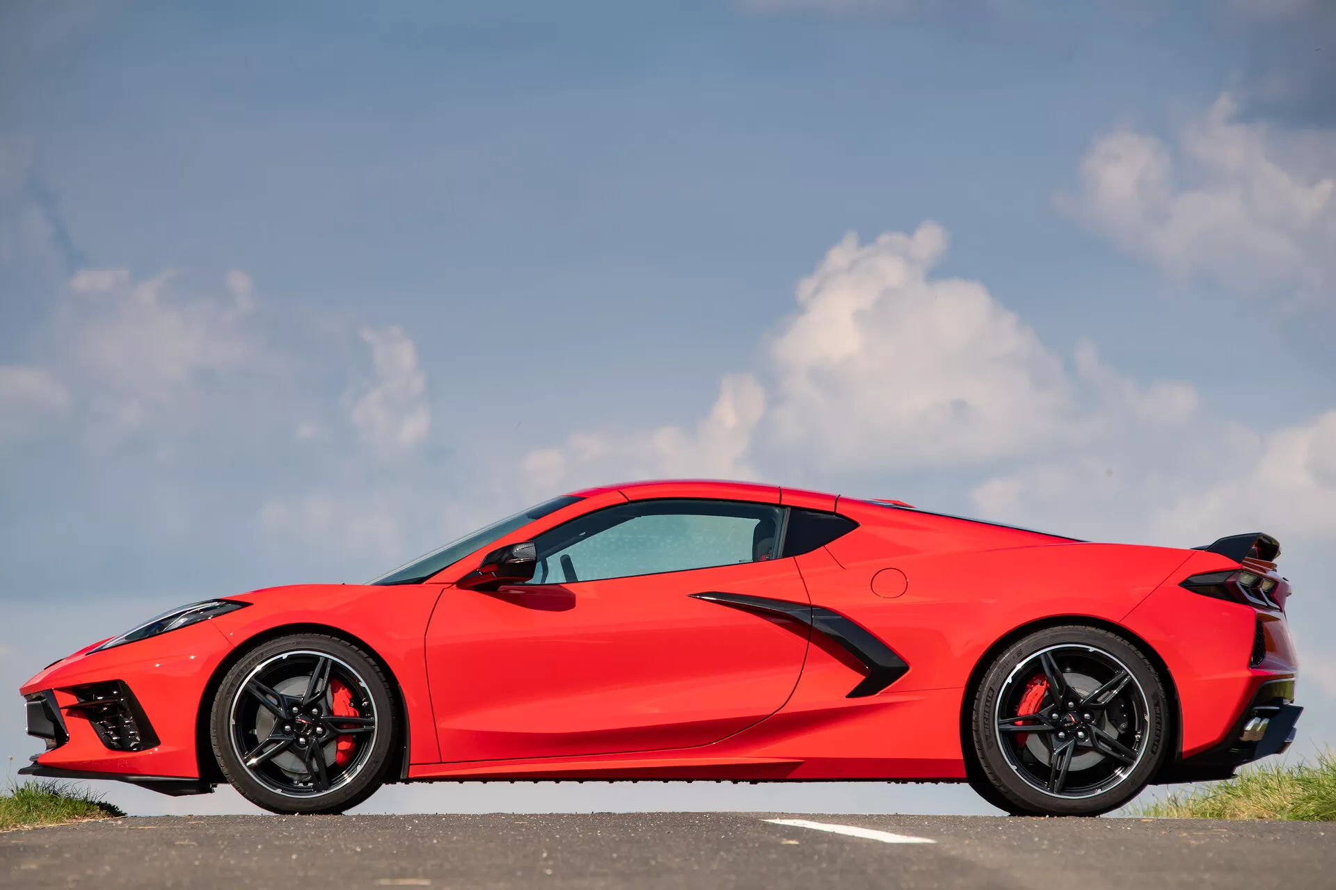 Stranski profil svetlo rdečega športnega avtomobila Chevrolet Corvette C8 iz leta 2021 proti modremu nebu z razpršenimi belimi oblaki. Avto, nameščen na cesti ali parkirišču, označenem z belo črto, ima eleganten aerodinamičen dizajn, ostre linije in krivulje. Izpostavljeno sprednje kolo in blatnik, črna večkraka lita platišča z nizkoprofilnimi pnevmatikami, stranska ogledala v barvi karoserije, velike zračne reže za vrati, zatemnjena okna, vgrajen ročaj vrat in zadnji spojler. Pogoji so jasni, idealni za fotografiranje, poudarjajo dizajn in estetiko avtomobila.