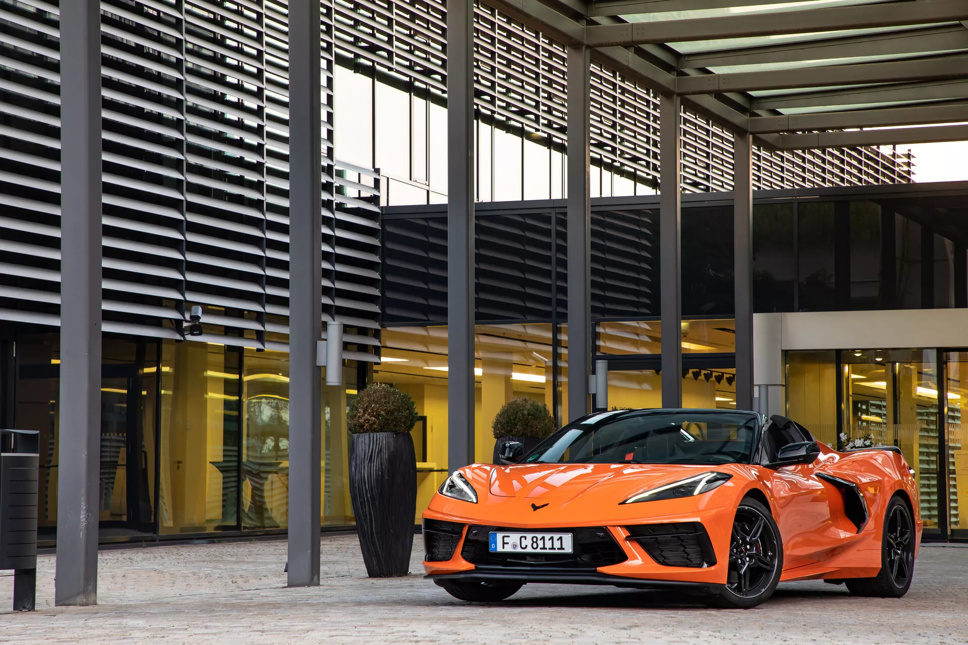 Športni oranžni kabriolet Chevrolet Corvette 2021 (C8) parkiran pred modernim stavbnim kompleksom s prepoznavnimi pravokotnimi vzorci tramov in stebrov; tričetrtinski pogled od spredaj, ki poudarja voznikovo stran, elegantne žaromete, dnevne LED luči, nizkoprofilne pnevmatike na večkrakih aluminijastih platiščih in aerodinamične oblikovalske poudarke. Odsevne površine prikazujejo sijajno barvno obdelavo in arhitekturno integracijo.