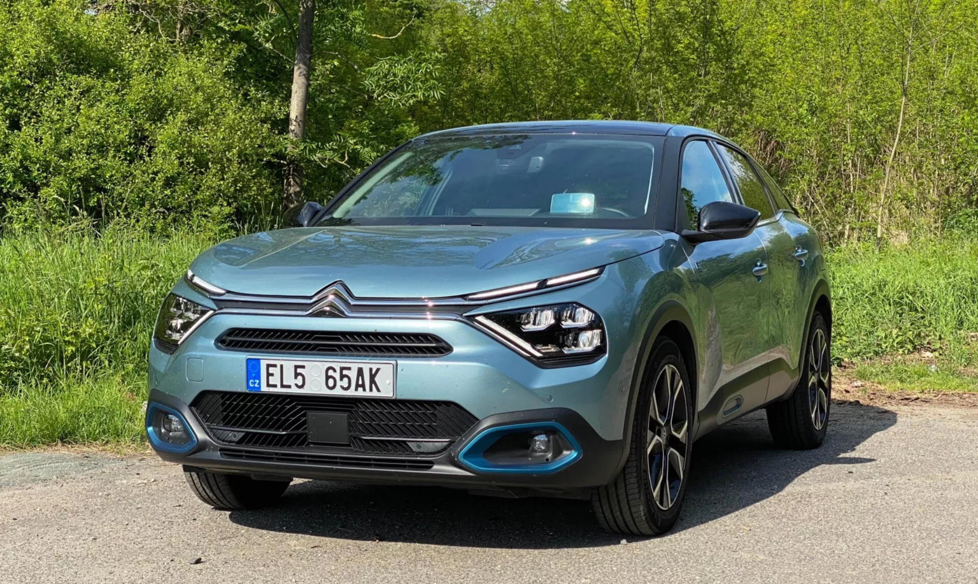 Slika sodobnega hatchbacka Citroen C4 (E-C4) 2020 iz nizkokotnega sprednjega tričetrtnega pogleda, ki prikazuje sprednji del, voznikovo stran, pokrov motorja, žaromete in dizajn koles. Značilnosti vključujejo LED dnevne luči okoli elegantnih enot žarometov, meglenke vgrajene v odbijač, centralno masko z logotipom proizvajalca, obkroženo z okrasno kromirano obrobo, gladke linije pokrova motorja, kljuke vrat in stranska ogledala, usklajena z barvo karoserije, več-kraka lita platišča, kovinsko barvo in sprednjo registrsko tablico. Avto je parkiran ob cesti z zelenjem v ozadju.