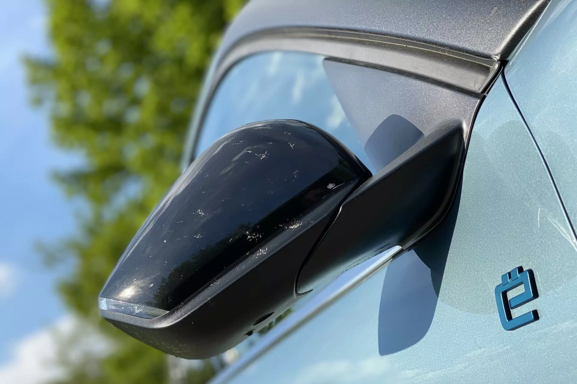 Pomorežni posnetek stranskega ogledala Citroen C4 2020 (E-C4), ki poudarja črno ohišje ogledala in kontrastno svetlo barvo karoserije. V ozadju je vidno jasno nebo in zelenje, kar kaže na zunanje okolje. Stanje avtomobila je novo, čeprav je na ohišju ogledala nekaj umazanije. Viden je tudi stiliziran emblem 'e', kar verjetno označuje električno različico.