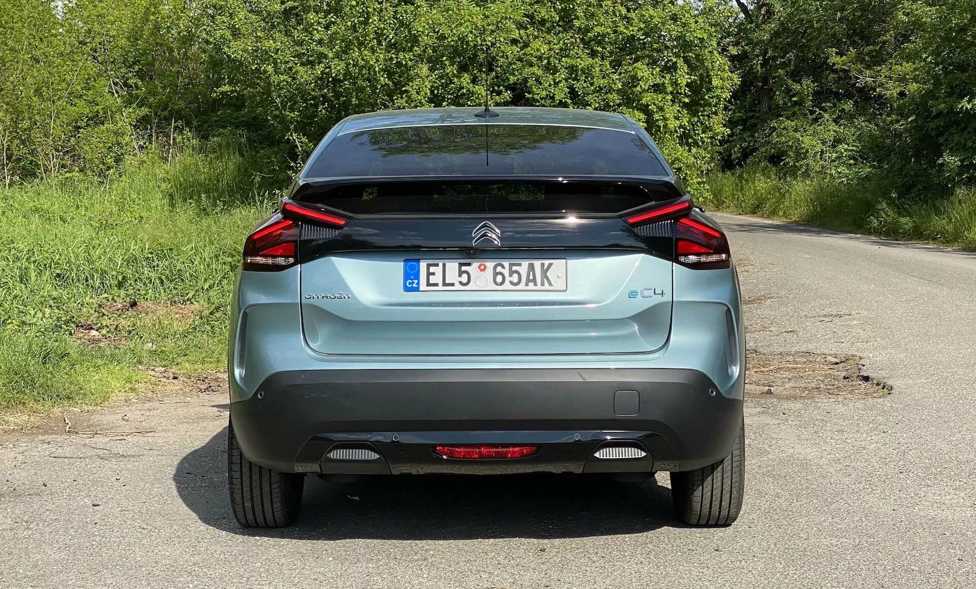 Zadnji pogled na modrega Citroena C4 iz leta 2020 (e-C4), parkiranega na podeželski cesti. Slika zajema celotno širino vozila od spodnjega dela koles do strehe, prikazuje značilne kotne zadnje LED luči, znak znamke, oznako 'C4', registrsko tablico v evropskem slogu, zadnji brisalec in črni spodnji del odbijača. Vidni so tudi odsevniki in zadnja meglenka.