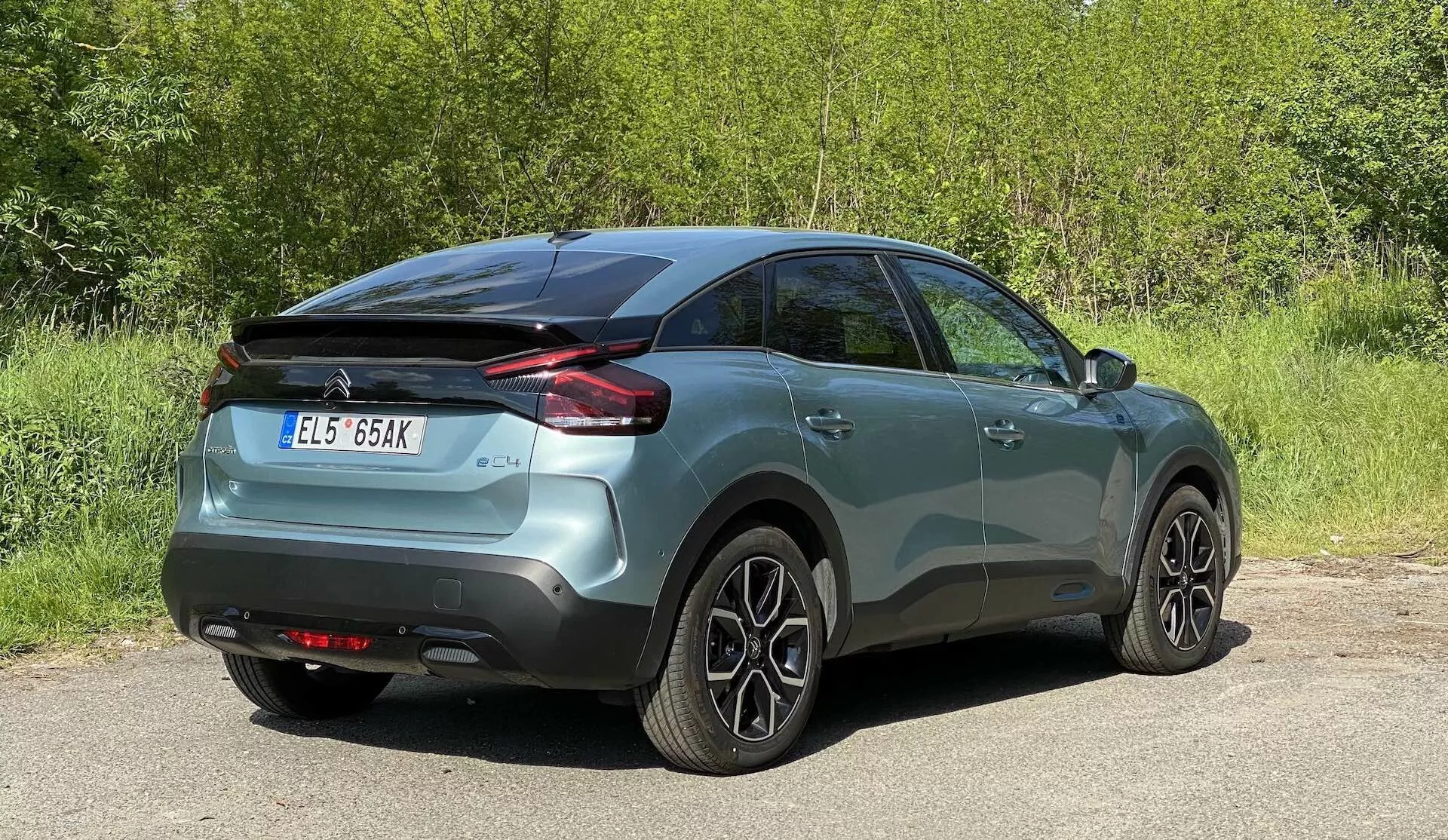 Slika avtomobila Citroen C4 2020 (e-C4), ki prikazuje zadnji triječetrtinski kot. Metallicno pobarvano vozilo je centralno postavljeno, razkriva bočne in zadnje profile, vključno s horizontalno zasnovanimi zadnjimi lučmi, barvnimi ročaji na vratih in večkrakimi aluminijastimi kolesi. Okolje nakazuje zunanjo nastavitev z nekaj rastlinja v ozadju.