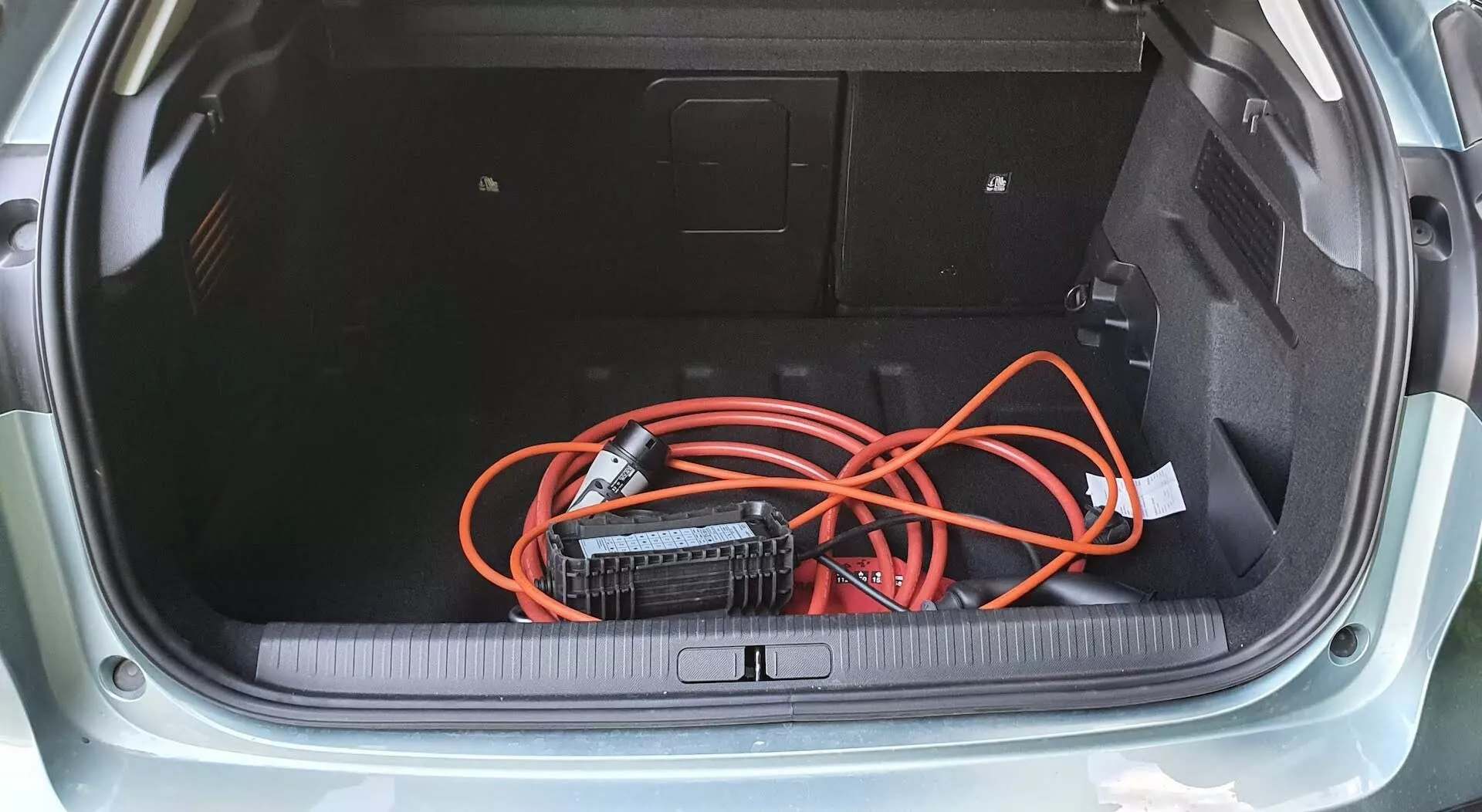 Pogled od zgoraj v prtljažnik kombilimuzine Citroen C4 2020 (e-C4) z preprogo obloženim dnom, stranskimi ploščami z dostopno ploščo in ohlapno postavljenim oranžnim podaljškom, priključenim na prenosni inverter. Prtljažni prag ima zaščitni pokrov iz gume ali plastike, kar poudarja praktično zasnovo shranjevanja vozila.
