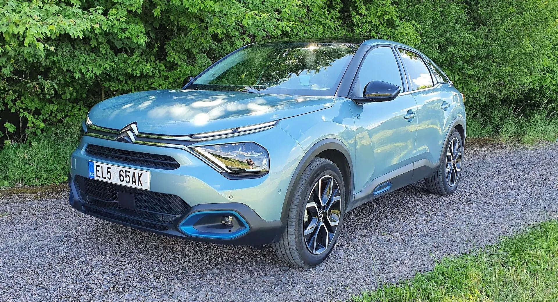 Svetlo modri Citroen C4 2020 (e-C4) kompaktni SUV parkiran na gramozni površini z vegetacijo v ozadju, pogled sprednjega kota prikazuje voznikovo stran, sprednjo fasado, izklesano karoserijo, LED žaromete, večkraka alu platišča, prednjo rešetko in značko proizvajalca, poudarjajoč oblikovalske in tehnološke lastnosti.