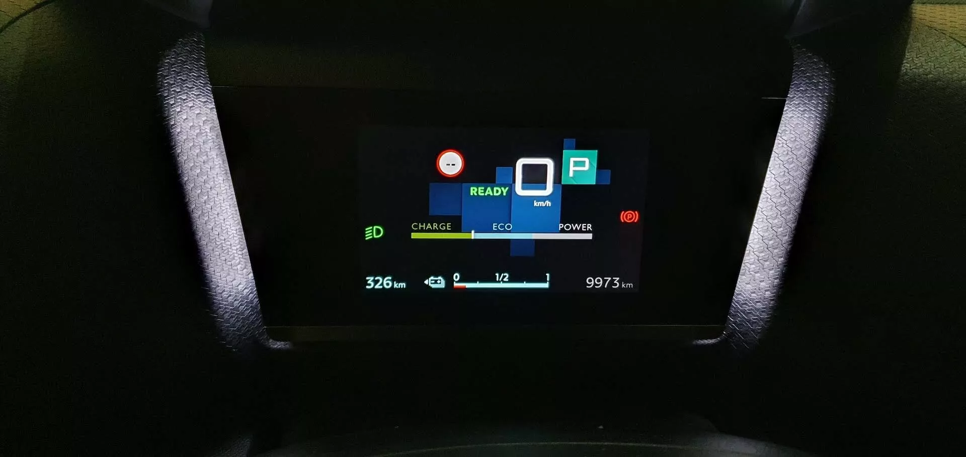 Odblizu pogled na digitalno nadzorno ploščo Citroen C4 2020 (e-C4), ki prikazuje indikatorje 'CHARGE' in 'ECO', signal 'READY', ikono baterije s '32.6', ikono ekološkega lista, indikator porabe 'POWER', simbol parkiranja 'P', '10' z navzdol obrnjeno puščico, ki kaže temperaturo, ter odčitek števca kilometrov '9973 km', zajet iz perspektive voznikovega sedeža.