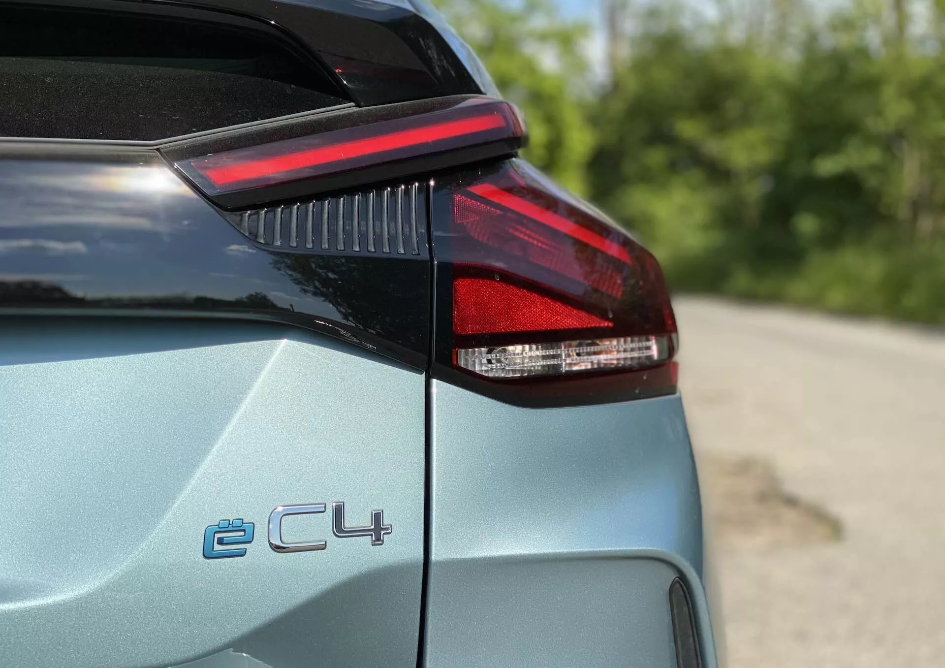 Podroben pogled na zadnji levi del Citroen C4 2020 (e-C4), prikazuje LED zadnjo luč, rob prtljažnika in del zadnje četrtne plošče; oznaka 'ë-C4', ki označuje električno različico; svetel kovinski odtenek z bleščečo površino; plitva globina ostrine, zamegljeno zunanje ozadje z zelenjem.