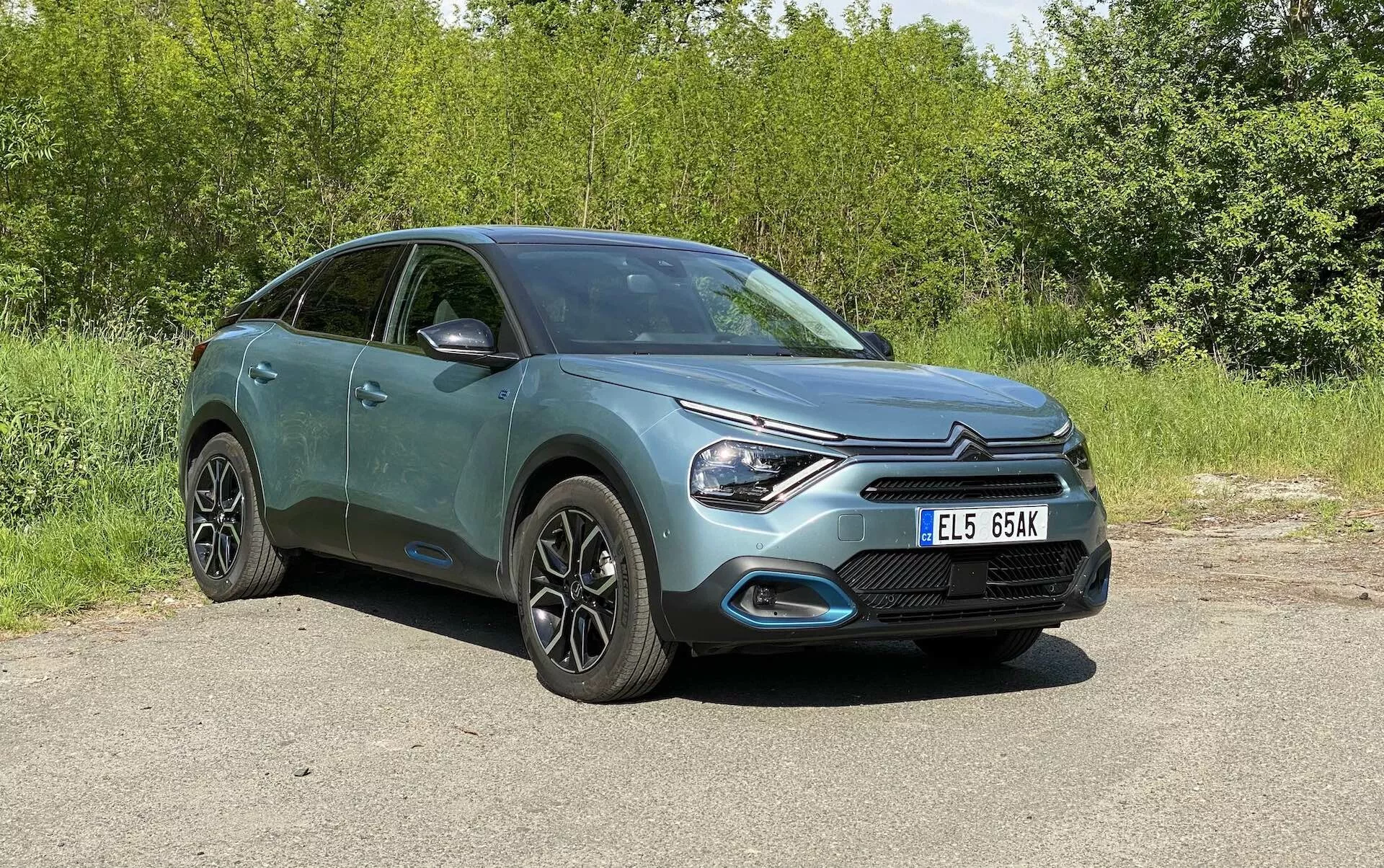 Celoten pogled na Citroën C4 2020 (e-C4) parkiran zunaj, posnet iz sprednjega tričetrtinskega kota, ki prikazuje sprednjo masko, eleganten dizajn LED žarometov in obliko pokrova motorja, skupaj z delnim stranskim profilom, vključno z vrati in stranskimi ogledali. Hatchback z elementi crossover/SUV oblikovanja ima moderna aluminijasta platišča, vgrajene smerne signale v ogledalih in kovinsko barvo. Ozadje je zeleno in avtomobil je prikazan na naravni dnevni svetlobi brez motečih elementov.