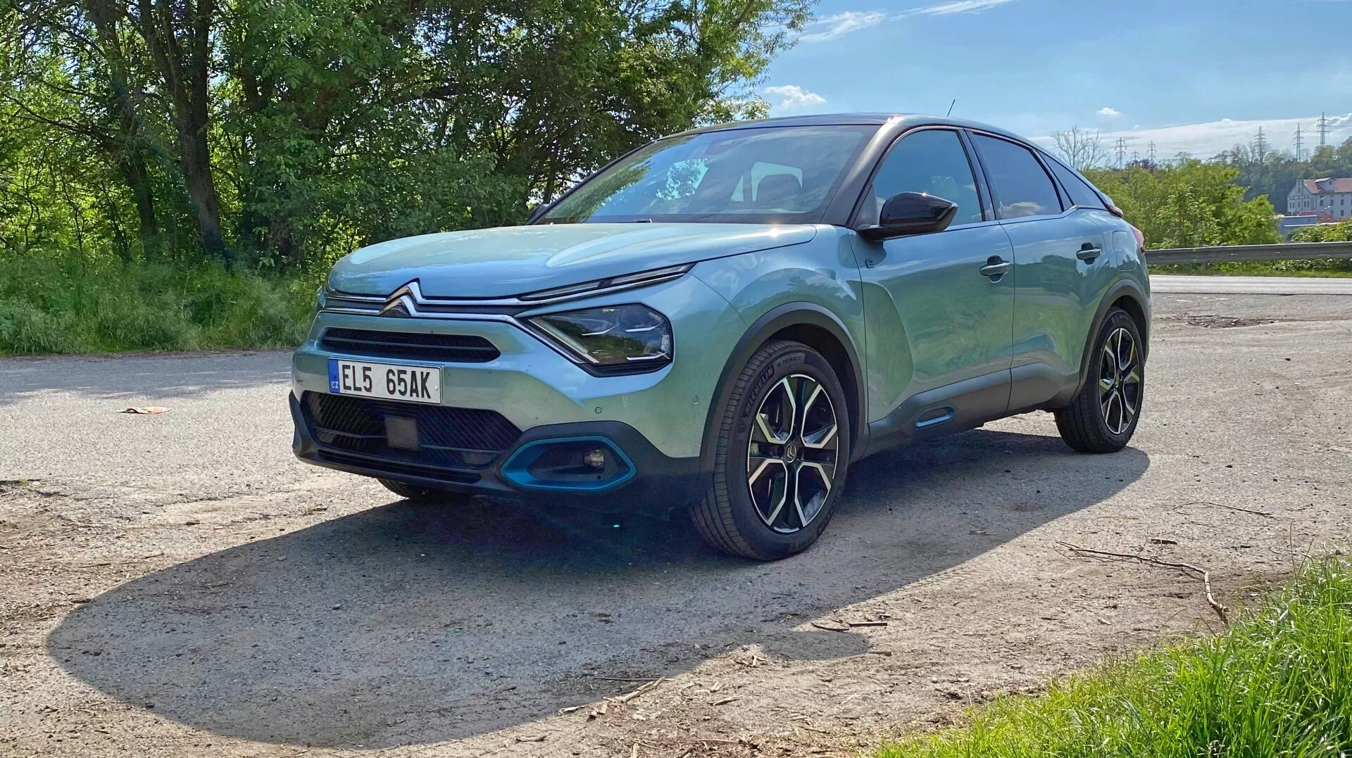 Slika prikazuje Citroen C4 (e-C4) hatchback ali kompaktni SUV iz leta 2020 iz nizkega prednjega trikotnega kota. Ta perspektiva poudarja sprednji del, vključno z rešetko, žarometi in odbijačem, ter voznikov profil, ki vključuje sprednji blatnik, kolo, voznikova vrata in del zadnjih vrat. Avto ima sodoben, športen dizajn z gladkimi linijami, nagnjeno streho, značilnimi LED sprednjimi žarometi in izrazito sprednjo masko. Prednji odbijač ima kontrastno črno oblogo, ki poudarja njegov robusten videz. Zapleten dizajn koles s številnimi kraki nakazuje poudarek na slogu ali zmogljivosti. Kovinska barva se v soncu svetleče blešči, kar odraža čist in poliran videz. Vozilo je parkirano na prostem v zelenem okolju, kot je razvidno iz dreves in jasnih vremenskih razmer. Tehnološke funkcije, kot so senzorji ali kamere, niso potrjene iz tega kota, registrska tablica pa je vidna, a za zasebnost izpuščena.