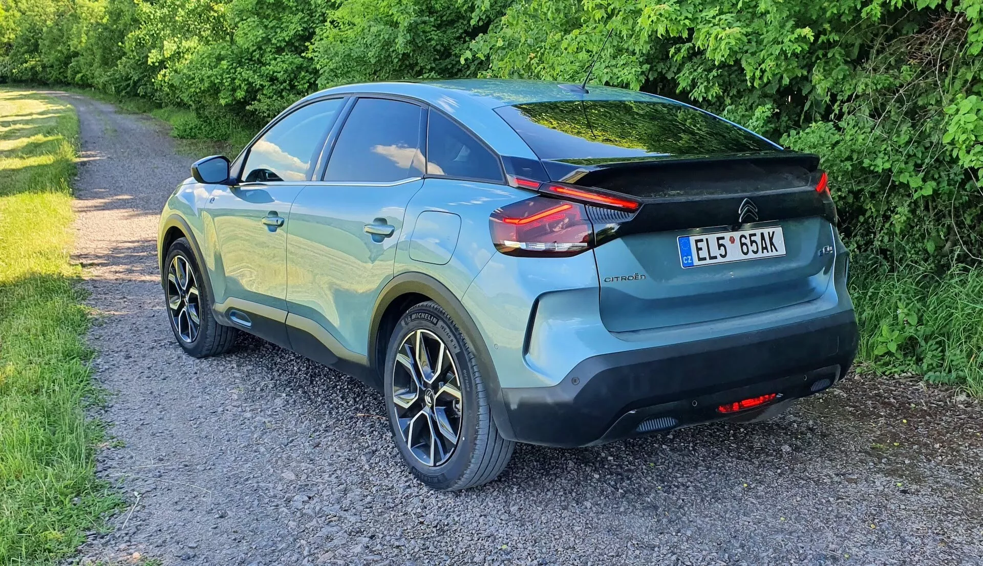 Slika prikazuje Citroen C4 2020 (e-C4) iz tristranskega pogleda, ki prikazuje sodoben dizajn crossoverja parkiranega na ozki poti z vegetacijo ob strani. Avto ima značilne LED zadnje luči, ki segajo v zadnje četrtinske plošče, strešni spojler, dvobarvno barvo in petkrake aluminijaste platišča. Vidni so zadnji brisalec vetrobranskega stekla, integrirani zadnji ročaji in reflektirajoči elementi na zadnjem odbijaču. Avto je v brezhibnem stanju, osvetlitev pa nakazuje pozno popoldne ali večer.