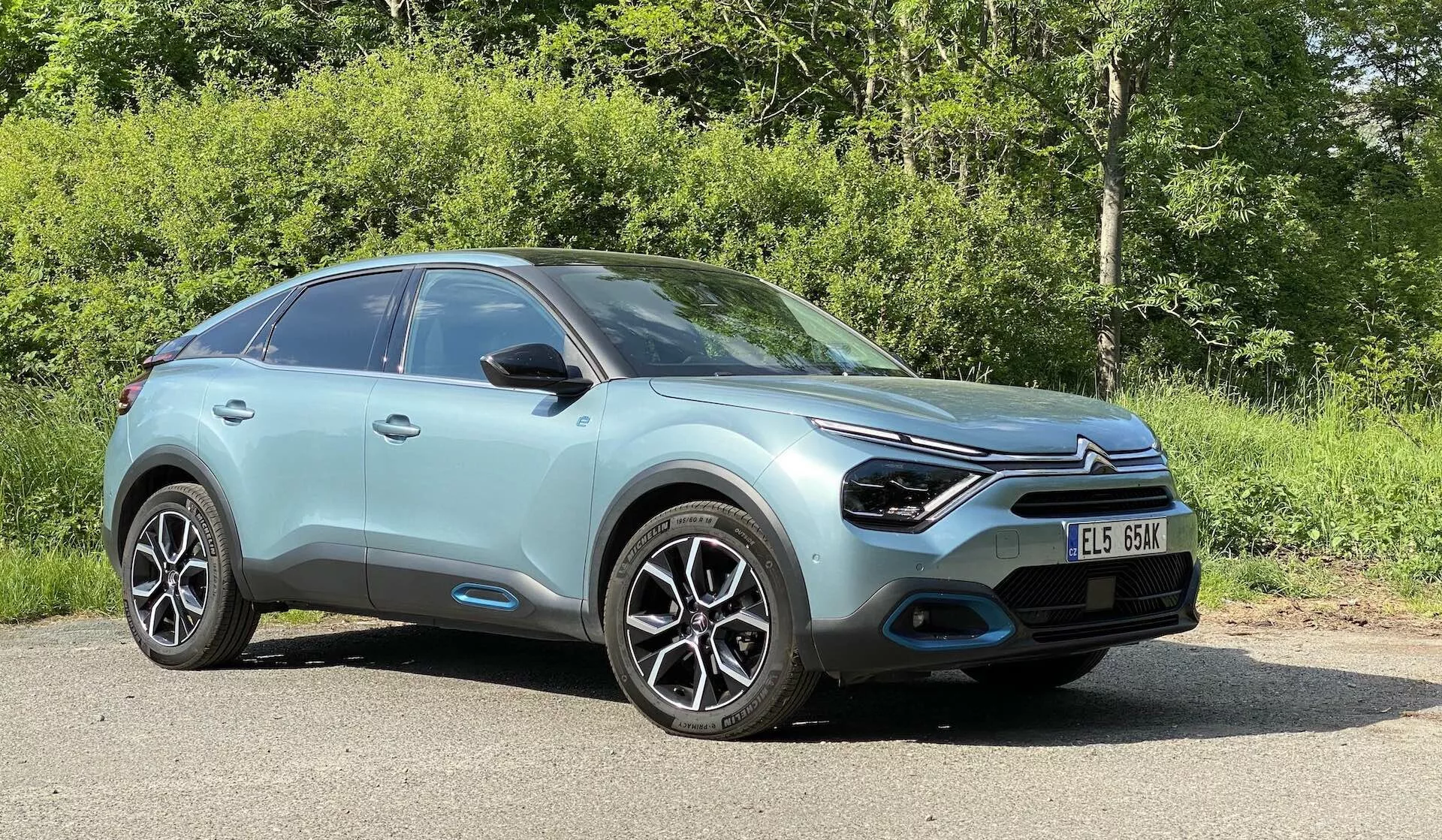 Nizek kotni pogled s strani Citroena C4 (e-C4) iz leta 2020, parkiranega na prostem, v naravnem okolju z vegetacijo v ozadju. Prikazuje eleganten, aerodinamičen dizajn, značilno sprednjo masko s centralno nameščenim emblemom, ozke LED žaromete, integrirane meglenke v črnih vložkih, večkraka lita platišča, sijajno mint ali svetlo turkizno barvo karoserije, gladke konturirane linije na pokrovu motorja in stranskih panelih, vzratna ogledala z integriranimi smerniki, rahlo nagnjeno strešno linijo, ki namiguje na karoserijski slog crossover SUV, črne obloge okoli kolesnih lokov in vzdolž spodnjih strani avtomobila ter del vetrobranskega stekla, ki odseva svetlo nebo na sončen dan.
