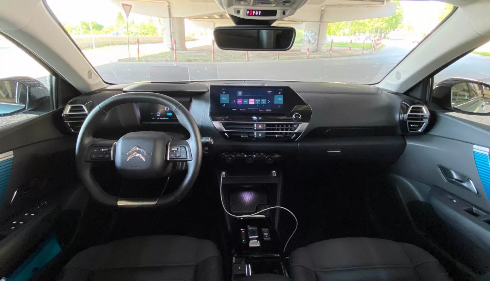 Pogled na notranjost Citroena C4 2020 (e-C4) iz zadnjega sedeža, osredotočen na armaturno ploščo z digitalnim instrumentom, sodobnim volanom z integriranimi kontrolami in vidnim zaslonom za informacijsko-zabavni sistem na sredinski konzoli. Konzola vključuje gumbe za nadzor klime, prestavno ročico in dodatne kontrole, kar nakazuje funkcije, kot so GPS navigacija, multimedijski sistem in ročni ali avtomatski klimatski nadzor. Prednji sedeži se zdijo oblazinjeni z vzglavniki, vratne plošče pa imajo premium poudarke.