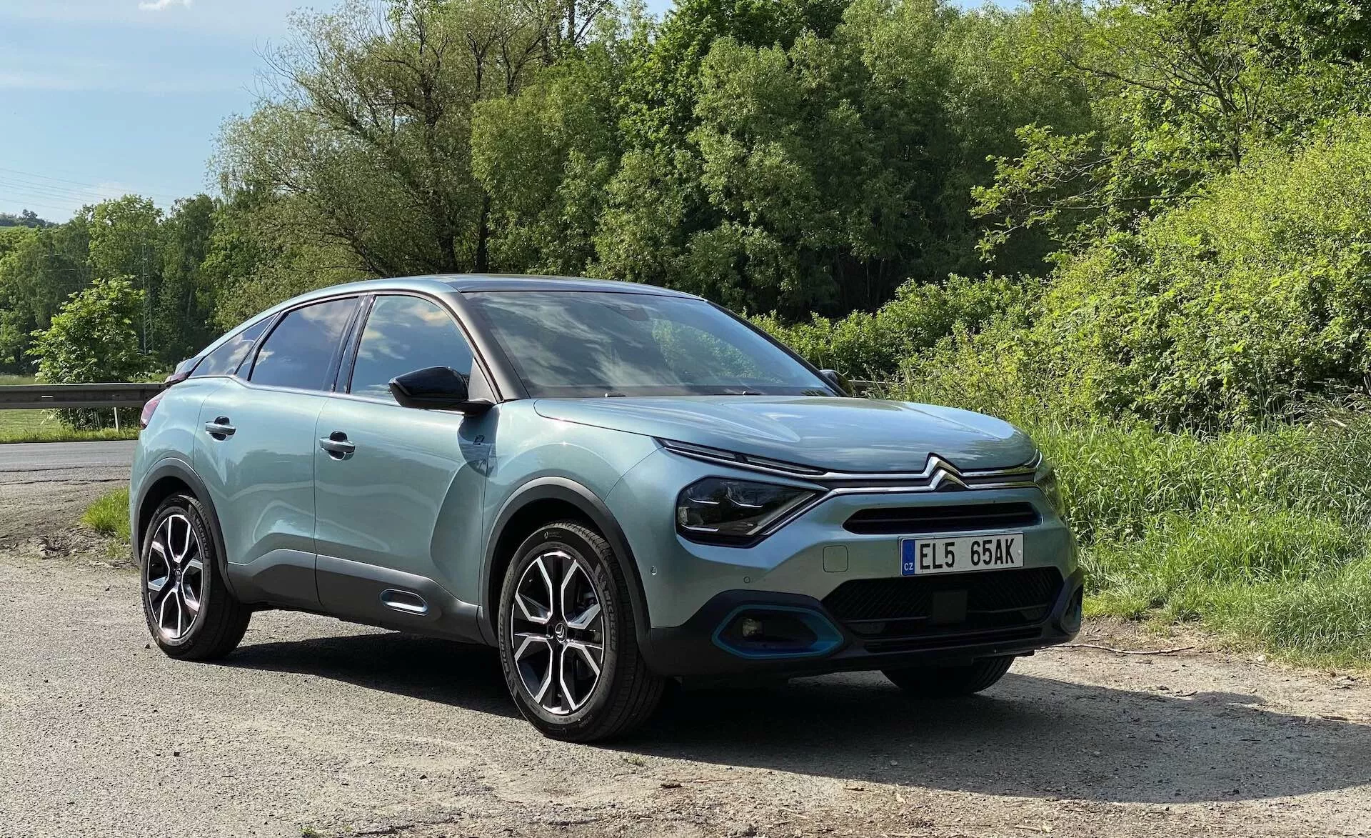 Slika Citroena C4 2020 (e-C4), posneta iz tričetrtinske čelne perspektive, poudarja pastelno modro karoserijo s kontrastno črno streho, sodobnimi LED žarometi, oblikovan prednji odbijač, lita platišča in zapleten vzorec naper. Slika, posneta iz rahlo navzdol usmerjenega kota, prikazuje podrobne poglede na levi žaromet, prednji odbijač, prednje kolo, vetrobransko steklo in voznikov stranski okni, s jasno vidno registrsko tablico. Avto je postavljen v zeleno okolico, bodisi podeželjsko bodisi parkovno, pod sončno dnevno svetlobo.