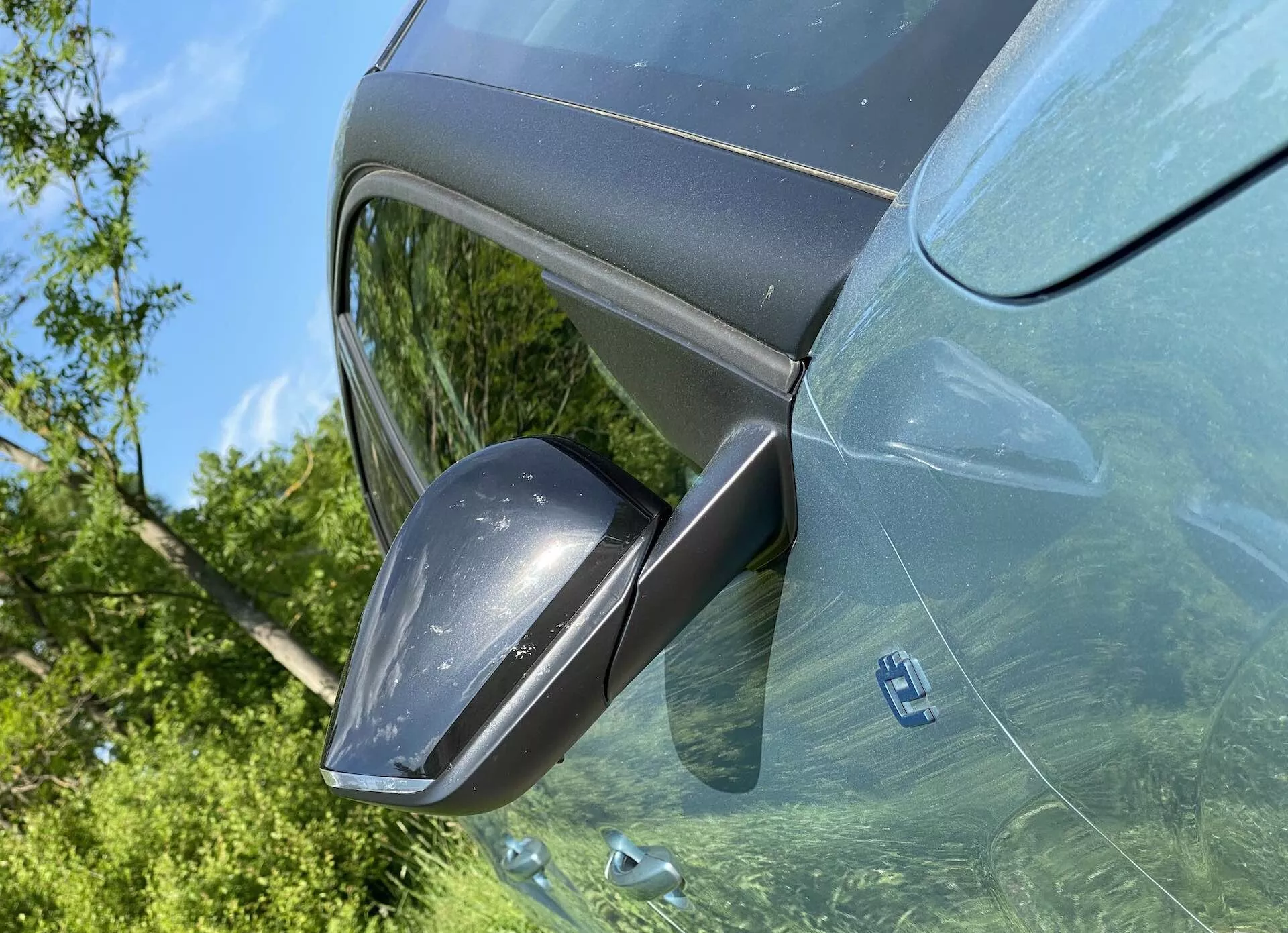 Povečava voznikove strani Citroena C4 2020 (e-C4) videna iz zgornjega kota, s poudarkom na sodobnem stranskem vzvratnem ogledalu z vgrajenimi smerniki, ohišjem, barvno usklajenim s karoserijo, in odsevajočem zunanje rastlinje na zatemnjenih oknih. Svetla dnevna scena poudari kovinski premaz in eleganten, sodoben dizajn vozila.