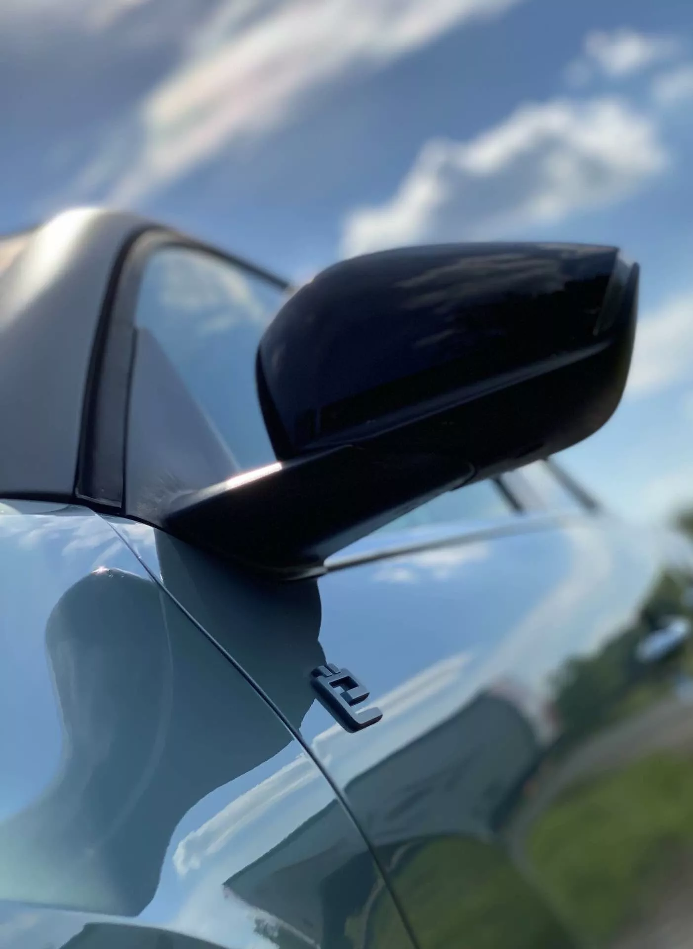 Povečan pogled na desno stransko ogledalo na sovoznikovi strani modela Citroen C4 2020 (e-C4) iz nizkega kota, ki prikazuje njegovo ohišje enake barve kot karoserija z črnim spodnjim delom v dvojnem barvnem oblikovanju. Poudarjeno je aerodinamično ohišje ter bleščeča površina in odsev neba z delno oblačnostjo pri naravni dnevni svetlobi. Del sprednjega blatnika in rob vrat razkriva sodobno oblikovanje avtomobila.