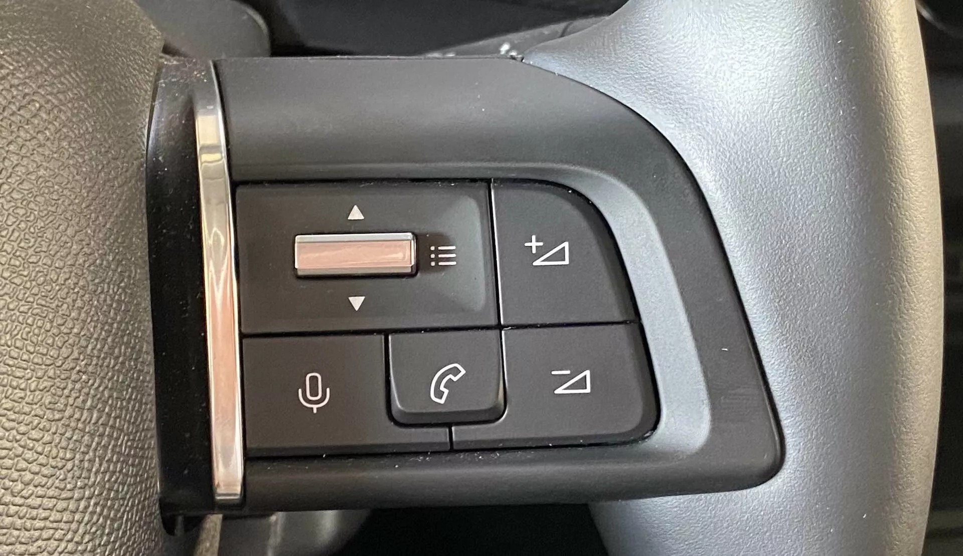 Povečan pogled na večnamensko nadzorno ploščo na levi strani volana v Citroënu C4 2020 (e-C4) iz perspektive voznika. Plošča vsebuje gumbe s simboli za uravnavanje glasnosti, preklapljanje skladb, upravljanje telefonskih klicev in joystick za navigacijo po meniju. Črna plošča in gumbi so zasnovani za taktilne povratne informacije in se ujemajo z estetiko volana, ki ima teksturirano površino in šiv, ki označuje visokokakovosten material.