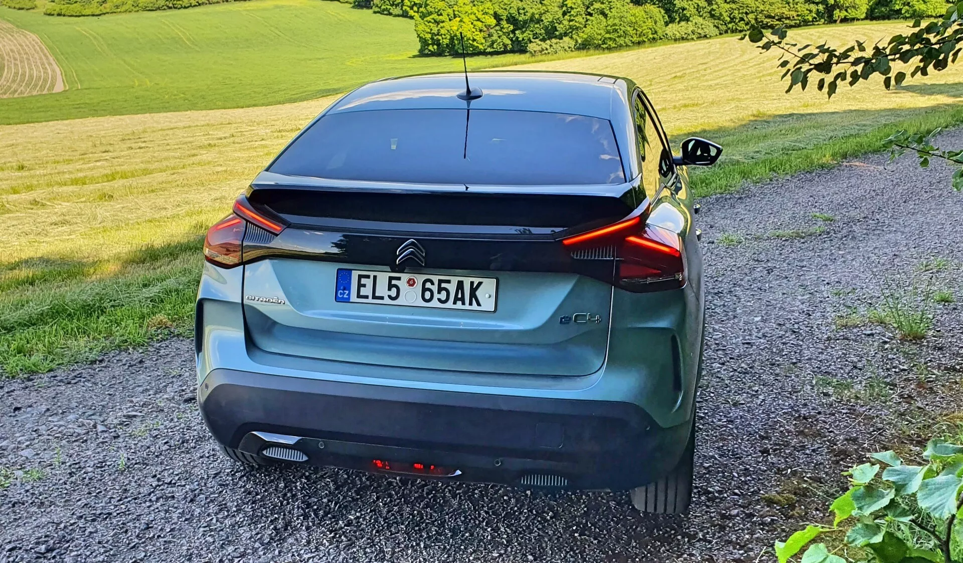 Zadnji pogled na sodoben hatchback avtomobil, ujet iz trikotnega kota, poudarja njegove elegantne linije, zadnje luči, sredinsko nameščeno registrsko tablico, aerodinamični spojler in zadnji brisalec. Vozilo, v sivem ali modrem kovinskem finišu, je opremljeno s senzorji na odbijaču. Avto je parkiran na makadamski poti v podeželskem okolju s travo in listjem, fotografirano pozno popoldne ali zgodaj zvečer z višine stoječe osebe.
