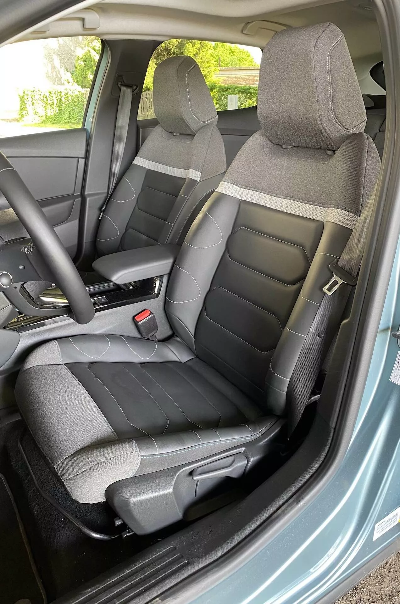 Notranji pogled Citroen C4 2020 (e-C4), osredotočen na voznikovo stran, posnet iz kota odprtih vrat, prikazuje volan, sedeže in del armaturne plošče z vgrajenimi krmilniki na volanu.
