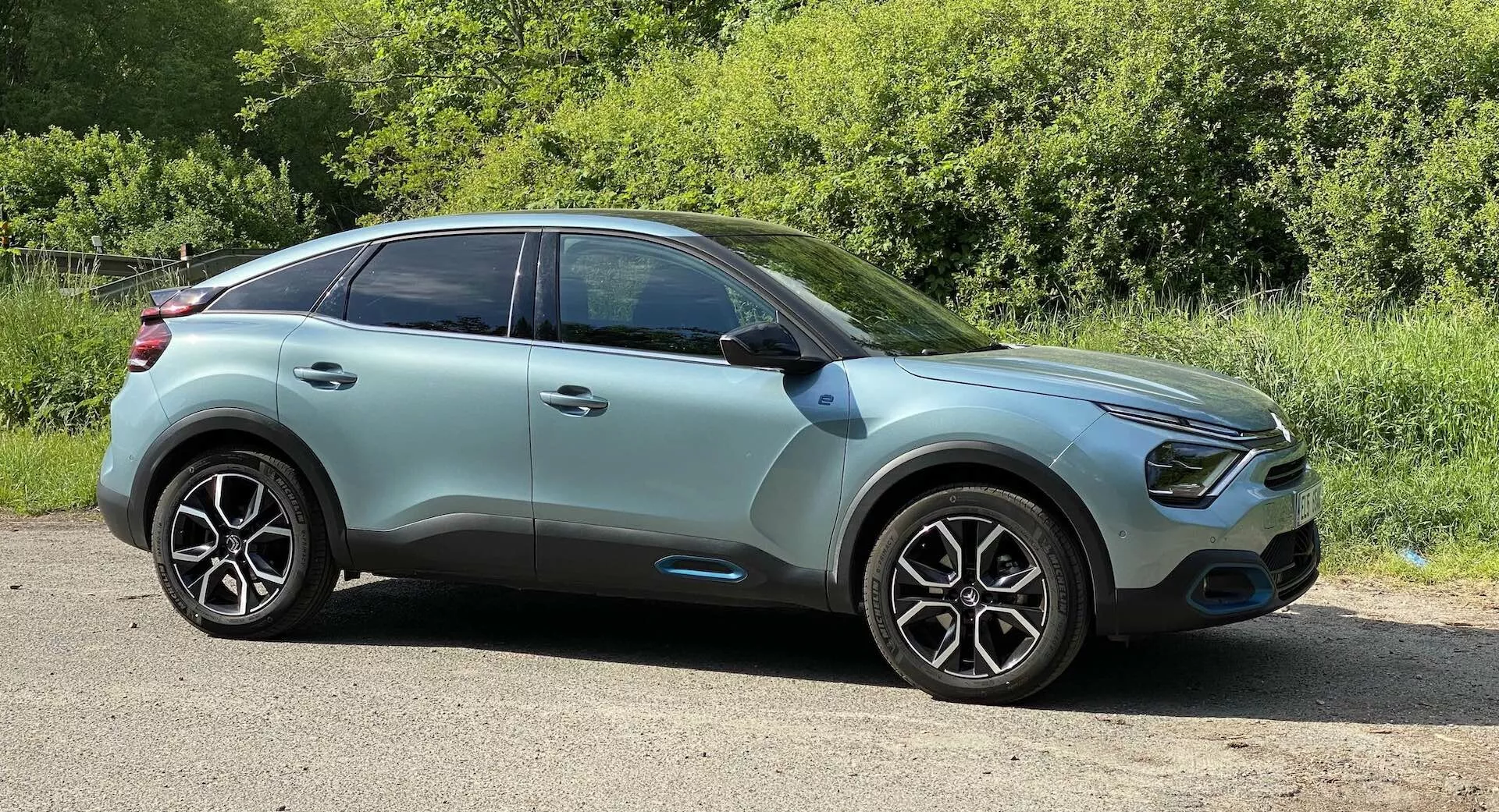 Bočni profil Citroen C4 2020 (e-C4) moderni crossover SUV posnet z nižjega kota. Avto je parkiran na ravni cestni površini s zelenjem v ozadju, kar kaže na podeželsko ali parkovno okolje. Elegantna pokrova motorja vodi do ukrivljene sprednje mreže, obkrožene z LED dnevnimi lučmi in potencialnimi meglenkami. Dvobarvna karoserija v svetlih turkizno zelenih barvah z črnimi obrobaki prikazuje napredne funkcije, kot so integrirane smerne luči v stranskih ogledalih in senzorji za brezključen vstop na barvno usklajenih kljukah. Velika, večkraka lita platišča in visoko zmogljive pnevmatike poudarjajo estetiko in zmogljivost. Nežno padajoča strešna linija, zatemnjena okna in potencialni napredni sistemi za pomoč vozniku nakazujejo osredotočenost na varnost, udobje in aerodinamiko. Posneto pri dnevni svetlobi z avtom, postavljenem na zmerni razdalji, slika poudarja zunanji dizajn vozila.