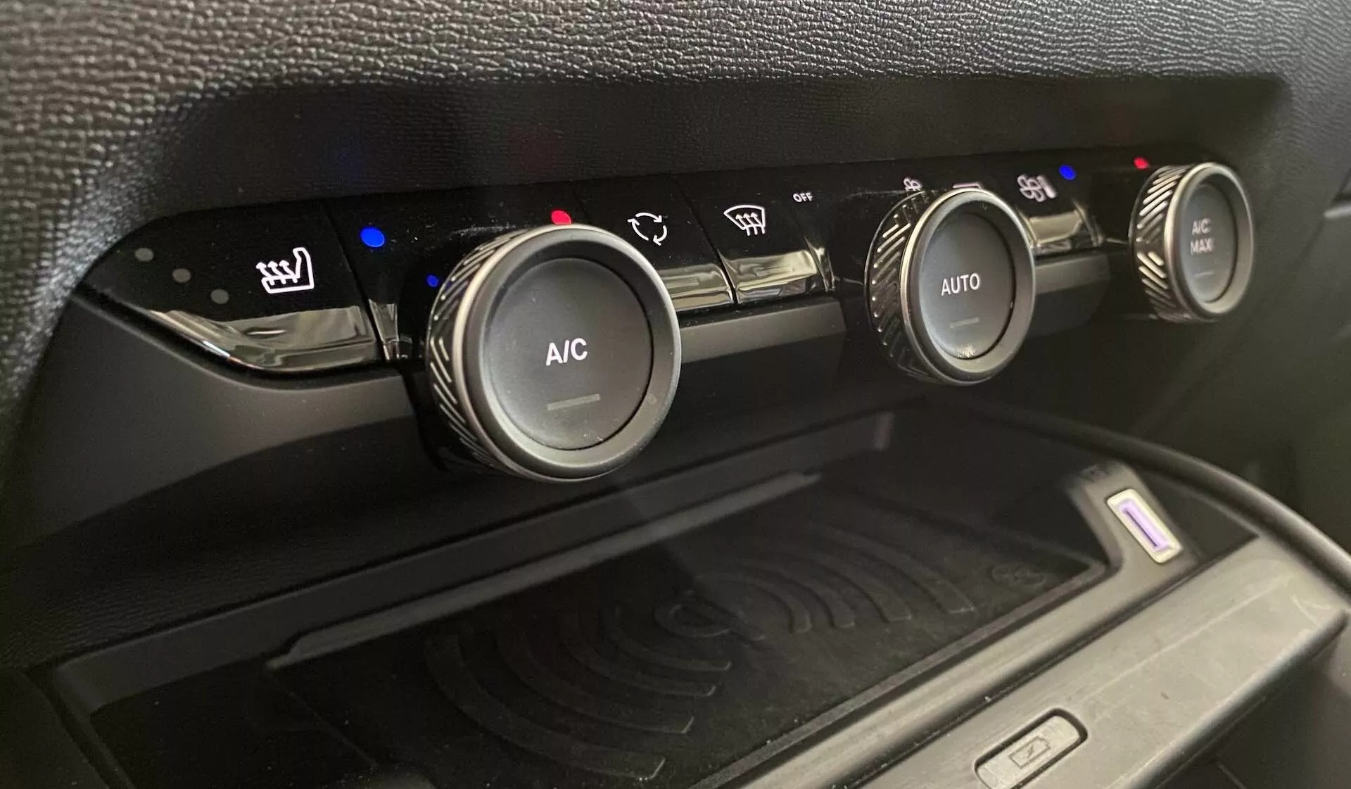 Pogled od blizu na klimatski sistem Citroena C4 2020 (e-C4), prikazuje tri krožne kontrolne gumbe na osrednji konzoli iz rahlo nagnjenega kotišča navzgor. Levi gumb nadzoruje ogrevane sedeže z modrimi indikatorji stopnje ogrevanja, srednji gumb je označen z 'A/C' za klimatizacijo, desni gumb pa ima 'AUTO' za samodejne nastavitve klimatske naprave. Modri in rdeči indikatorji odražajo nastavitve temperature. Pod gumbi se nahaja predel s podlogo za brezžično polnjenje z radijskim valovnim simbolom, obdan z mat črno plastično površino in teksturo z vzorcem.