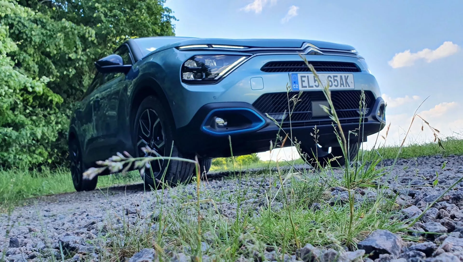 Nizek čelni pogled na parkiran Citroën C4 2020 (e-C4) na makadamski poti, obdani z zelenjem, ki prikazuje sprednji pokrov, mrežo, sprednji odbijač, žaromete in del sprednjih koles avtomobila. Slika poudarja robustno držo vozila in zunanje oblikovalske elemente, kot so oblika žarometov, vzorec mreže, zračni vhodi in linije pokrova motorja. Svetloba, ki se odbija od laka, poudarja estetsko privlačnost avtomobila. Možne napredne svetlobne tehnologije in parkirni senzorji so prav tako del vidnih tehnoloških funkcij.